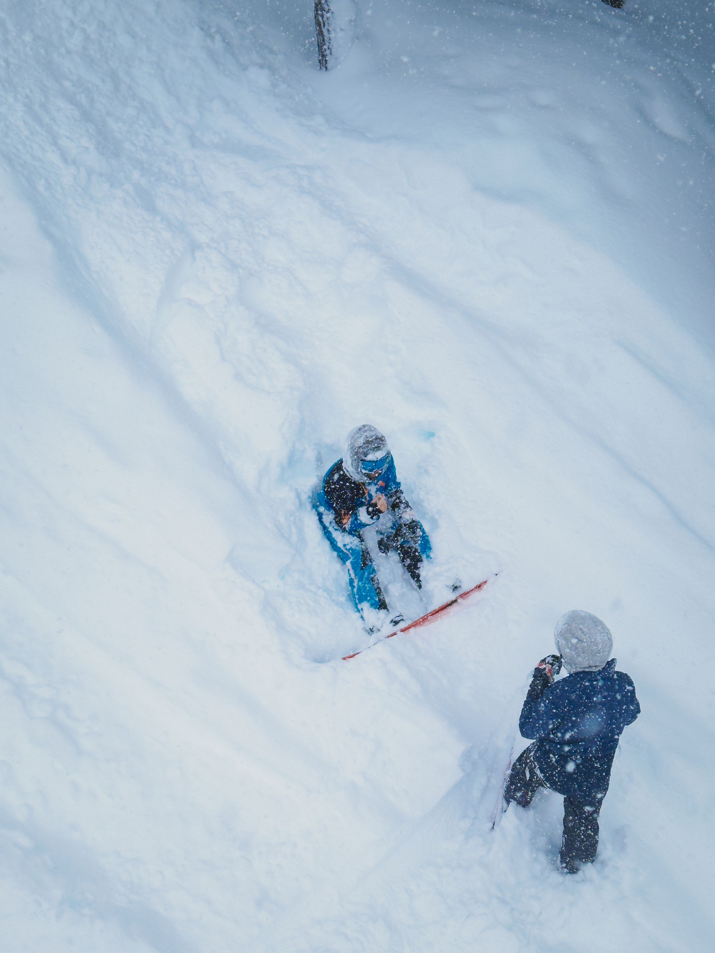Two children playing in deep snow; one is sitting on a snowmobile and the other, standing nearby, is dressed in winter clothing and interacting with the snowmobile.