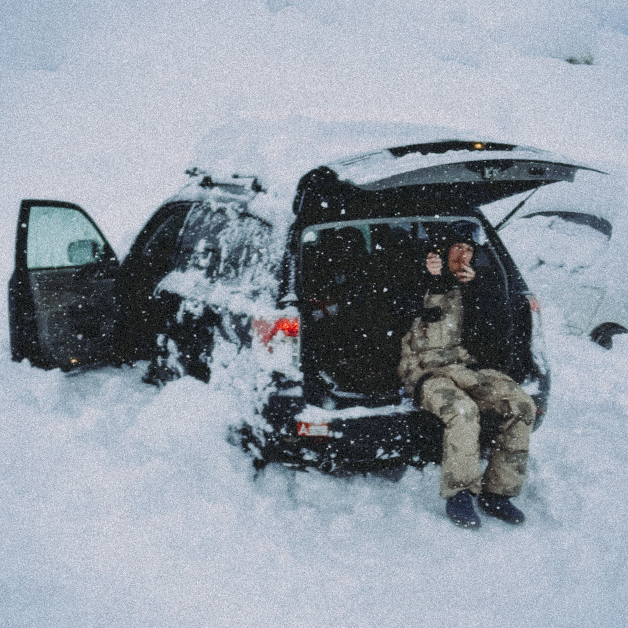 Person in camo sitting on the open trunk of a BLACK SUV in snowy landscape, with snowy weather and drifting snow.