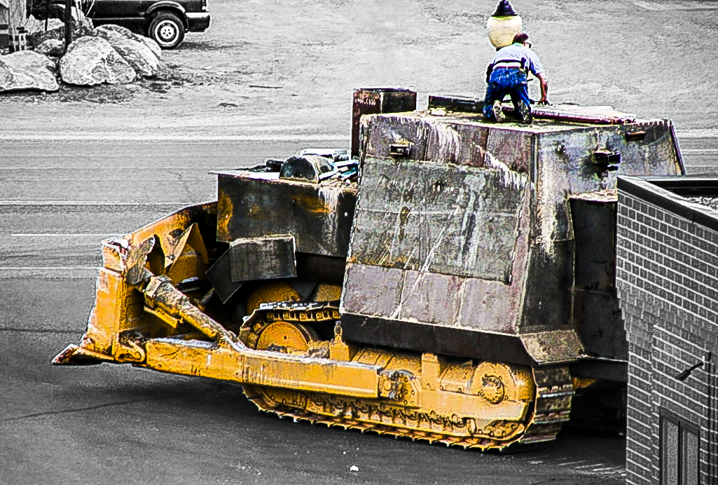 A large yellow and black construction vehicle, likely a bulldozer, is on a street, with a man in a blue outfit and hat standing on top of it. Killdozer.