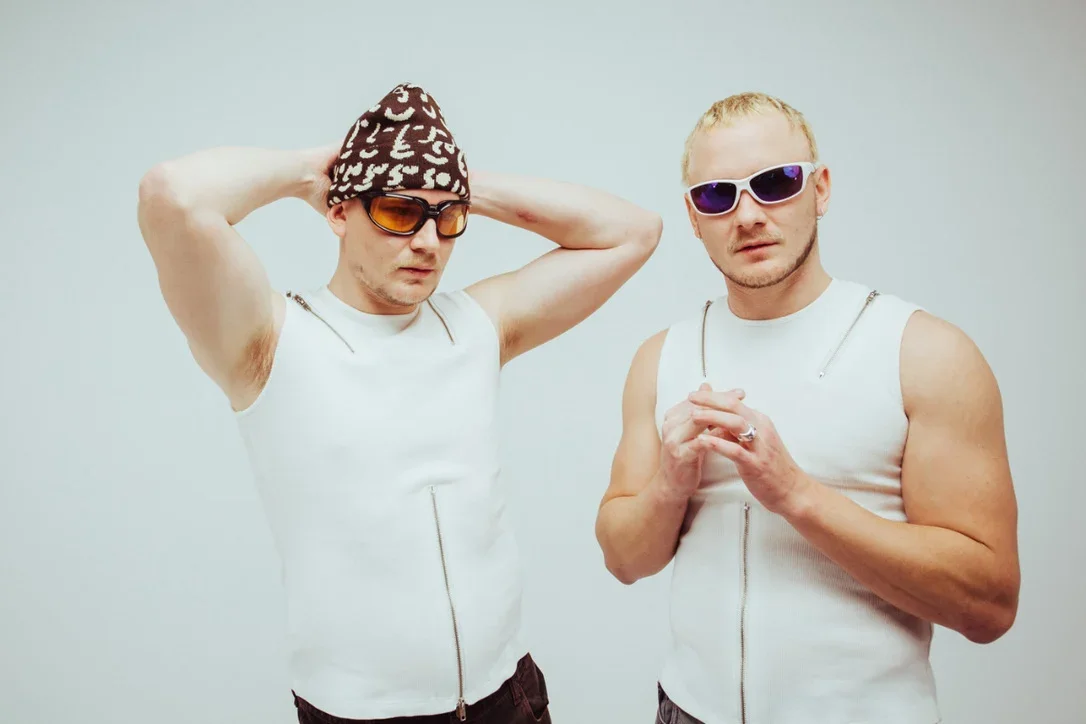 Cosmo's Midnight new press photos - Two men wearing sleeveless white tops with zipper details, sunglasses, one with a patterned hat, stand against a plain light background.