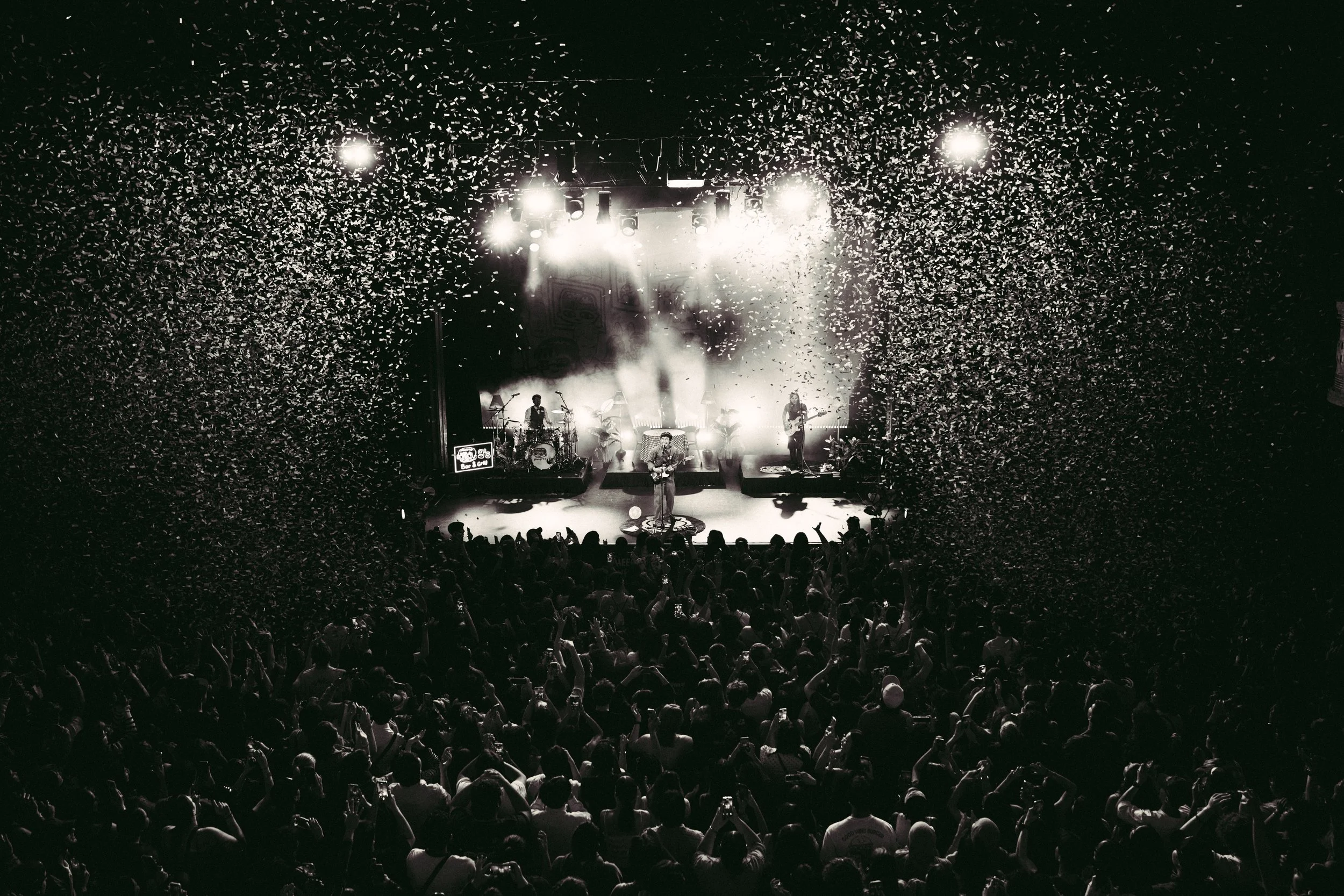 A live concert with grentperez and band performing on stage, confetti falling, and a large crowd dancing and taking photos.