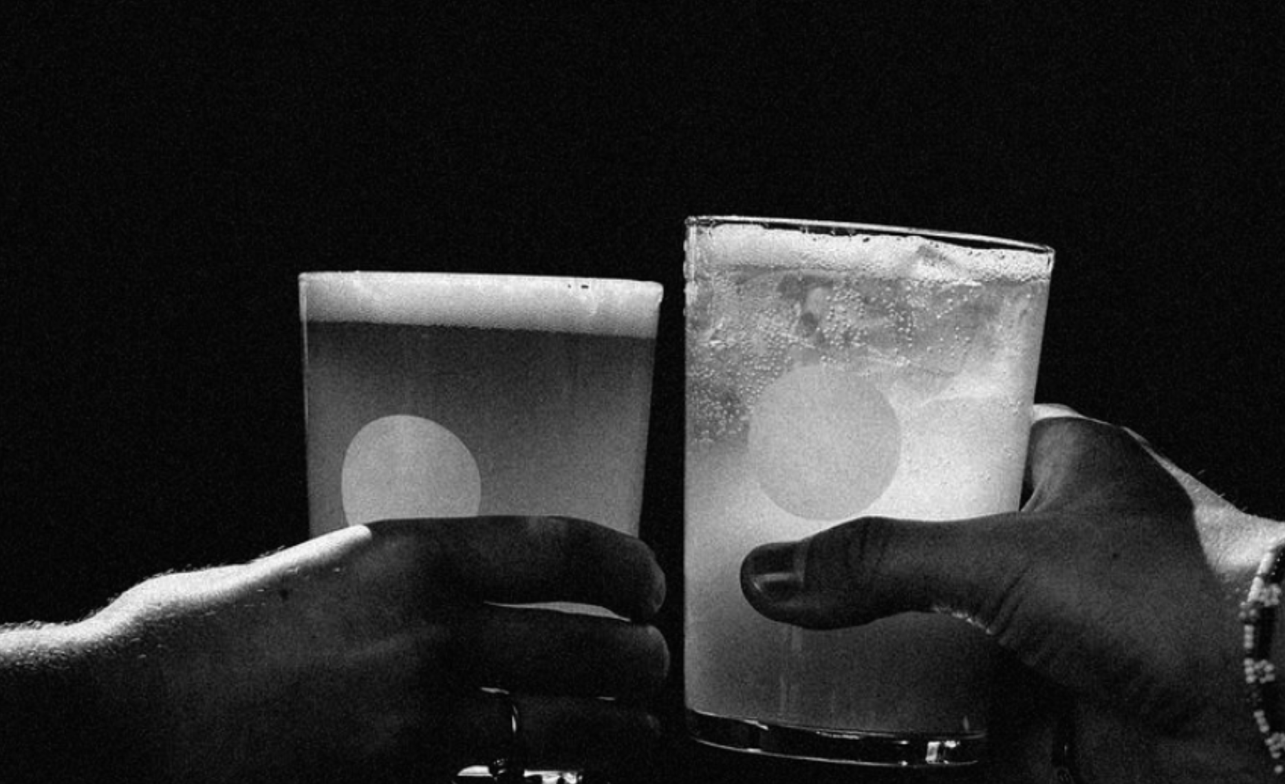 Two hands holding glasses of drink, one glass appears to contain a dark beverage, the other a clear, bubbly drink, against a dark background.