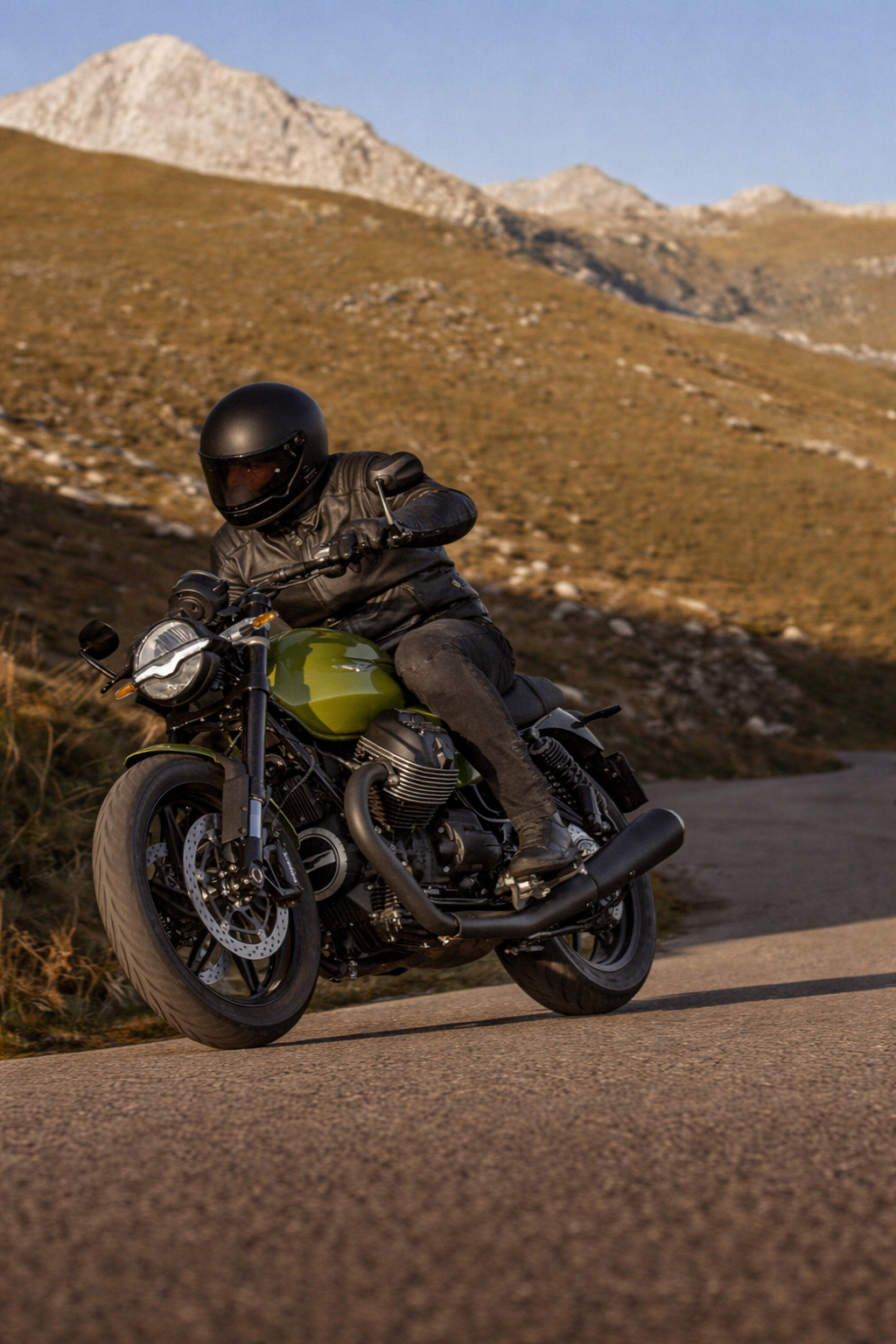 Motorcycle rider wearing a black helmet and leather jacket riding on a mountain road with a scenic background of mountains and clear sky.