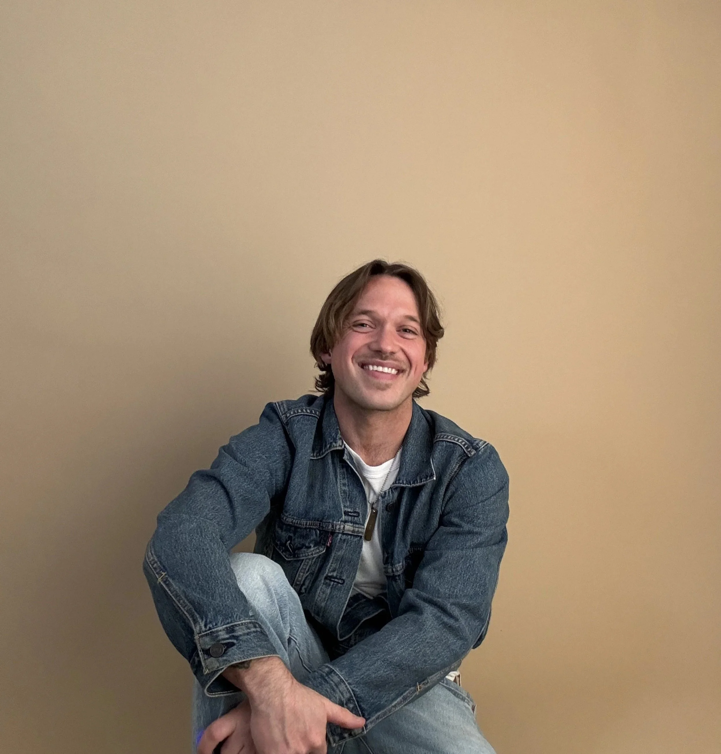 Smiling man with brown hair wearing a denim jacket and light jeans, sitting against a plain beige wall.
