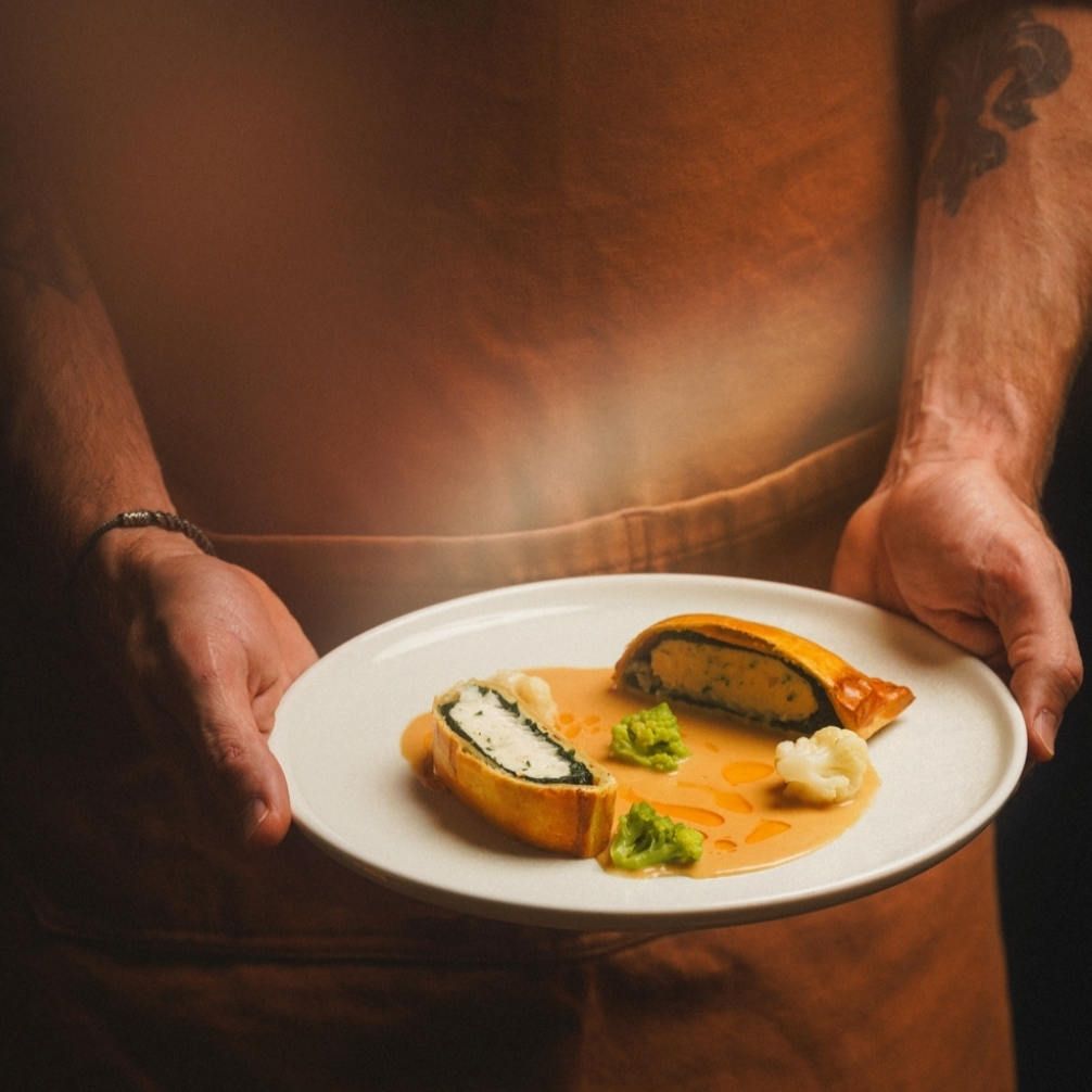 Person holding a white plate with a sliced stuffed vegetable, green sauce, and cauliflower