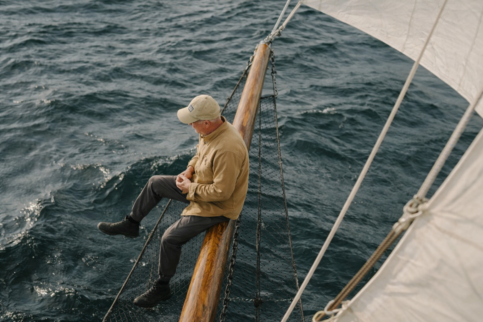 A man sitting on the edge of a boat's wooden railing over the water, with a sail visible on the right side of the image.