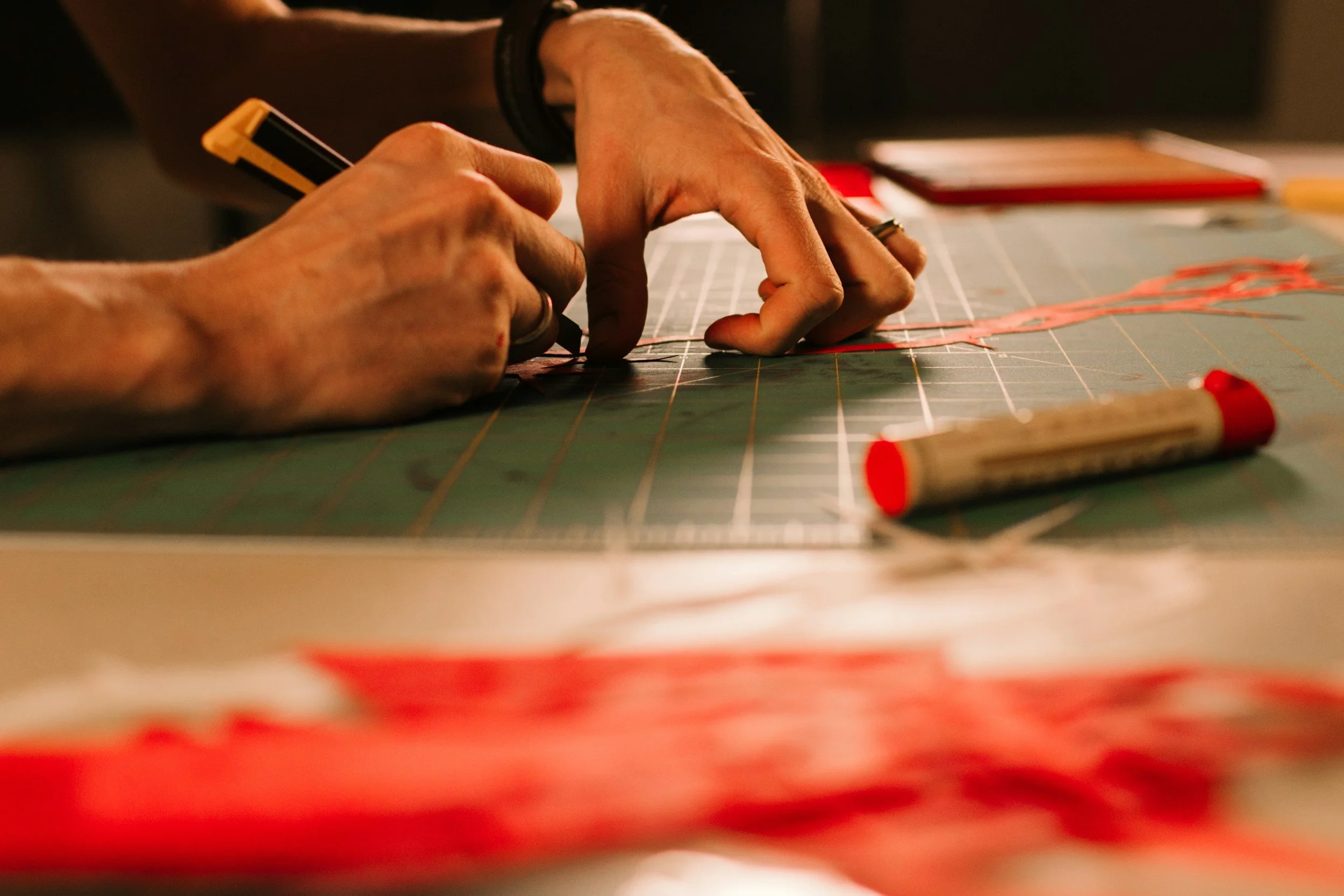 Close-up of hands using a ruler and a cutting tool on a green cutting mat with red and black details, likely working on a craft or design project.