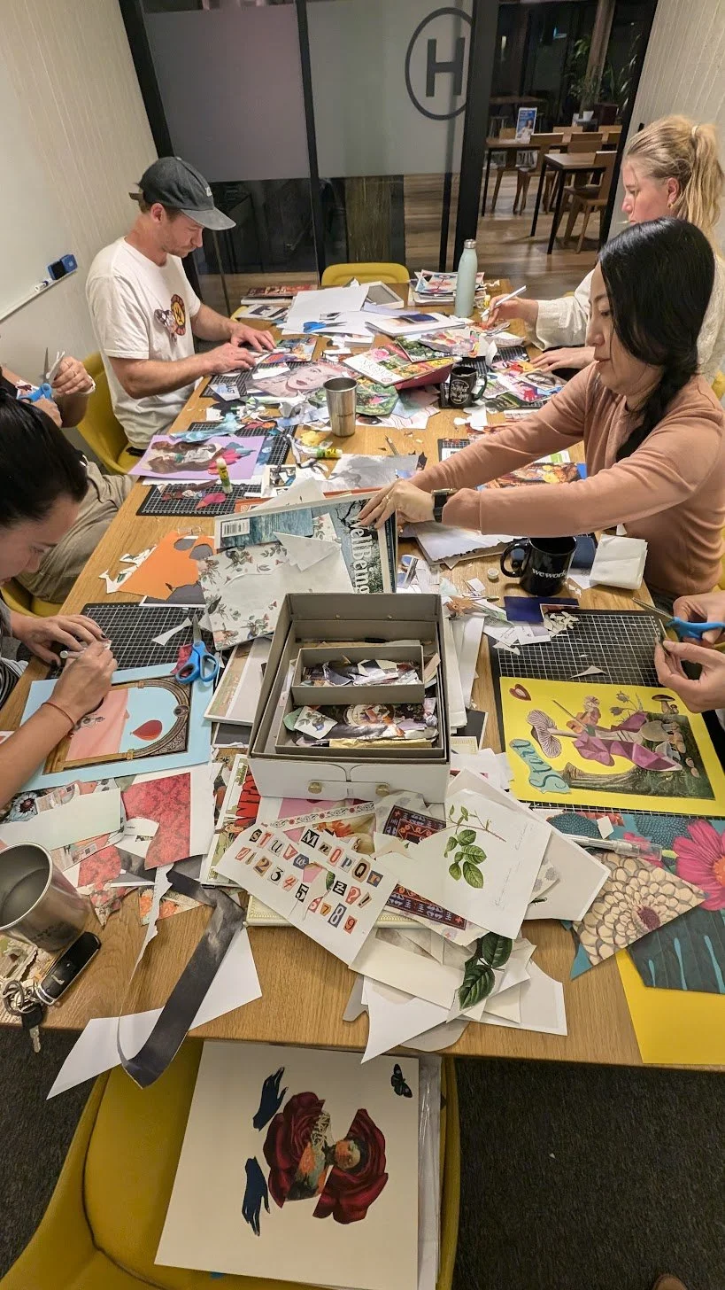 A group of six people working at a table covered with colorful paper scraps, scissors, and craft supplies, creating arts and crafts in a room with a glass wall and wooden furniture.