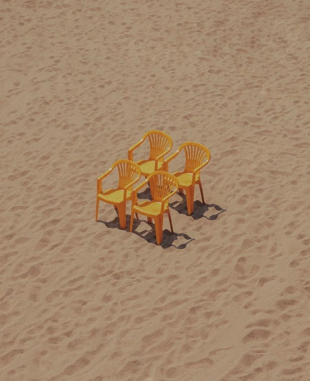Four yellow plastic chairs arranged in a square on sandy beach with visible footprints.