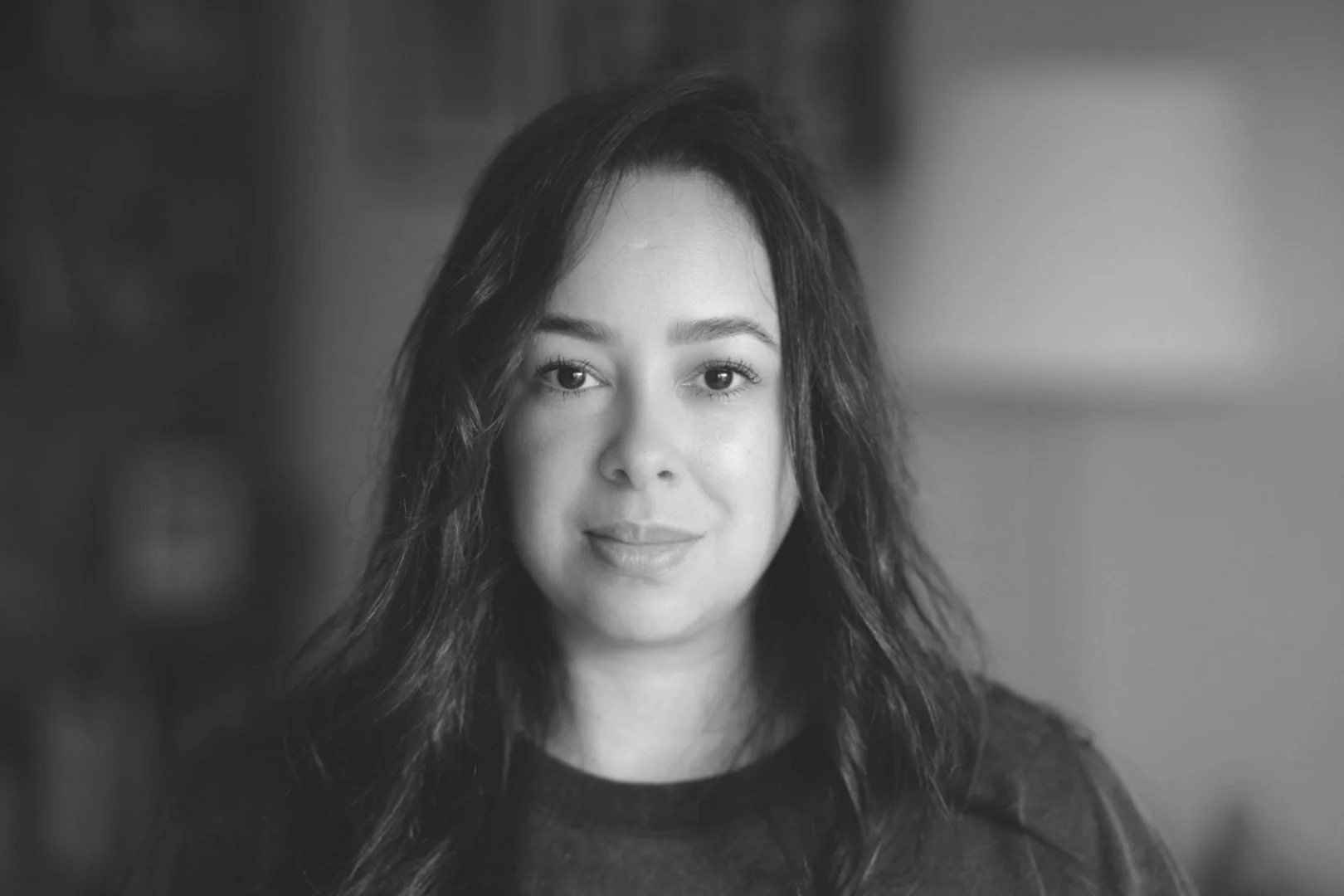 A black-and-white portrait of Sue, the founder and artist, featuring with long, dark, wavy hair, looking directly at the camera with a gentle expression.