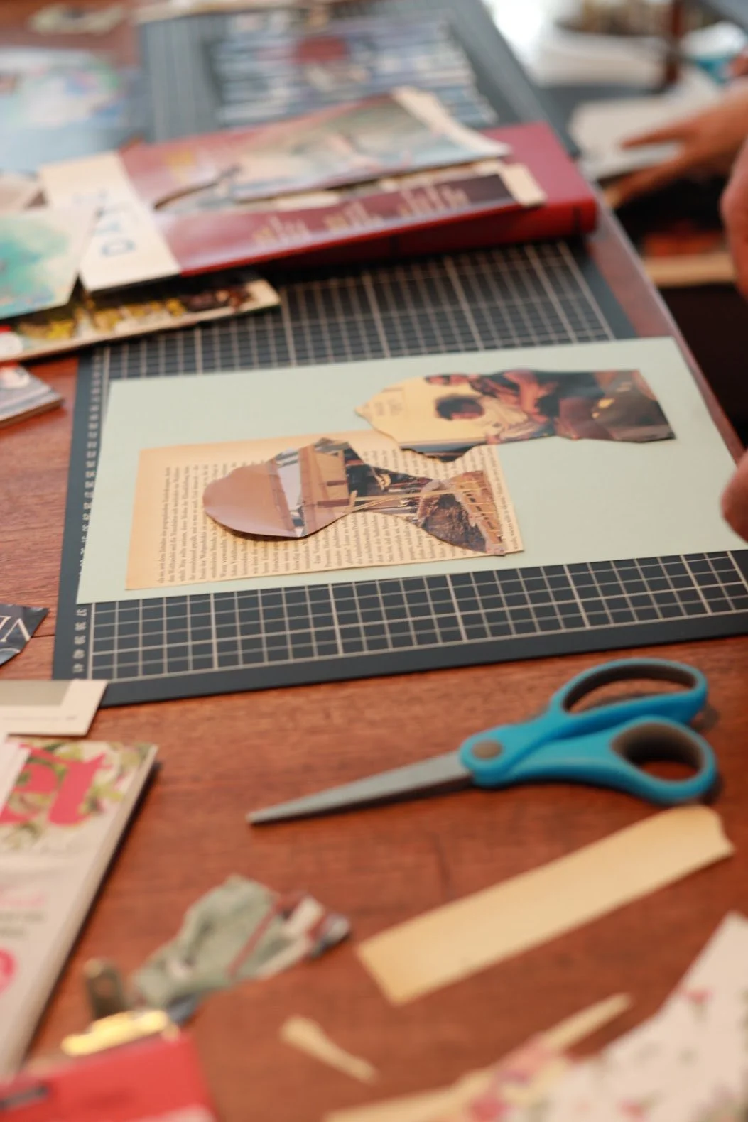 A desk with scrapbooking materials, including a pair of blue scissors, torn magazine pages, a cutting mat, and various cut-out images and paper pieces.