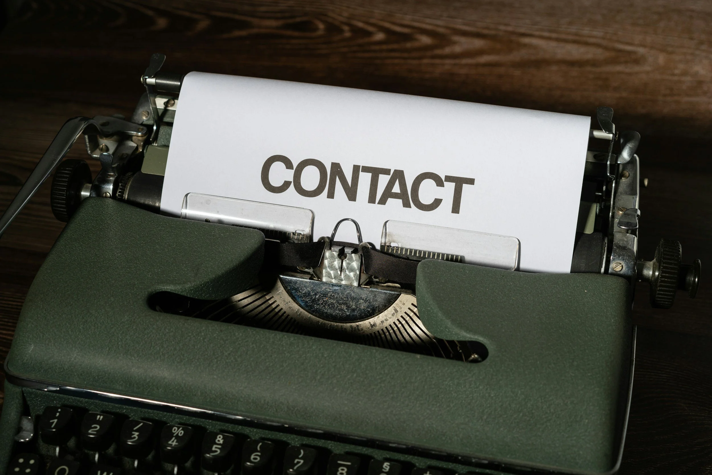 A vintage green typewriter with a sheet of paper that says 'CONTACT' on it, placed on a dark wooden surface.