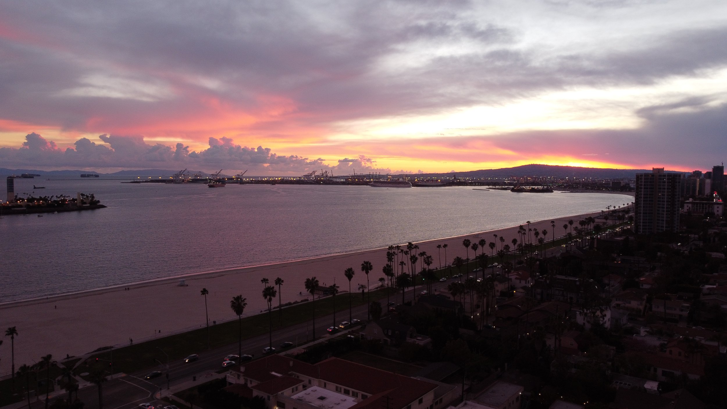 Sunset over a beach with palm trees, high-rise buildings, and a body of water, with clouds and a colorful sky.