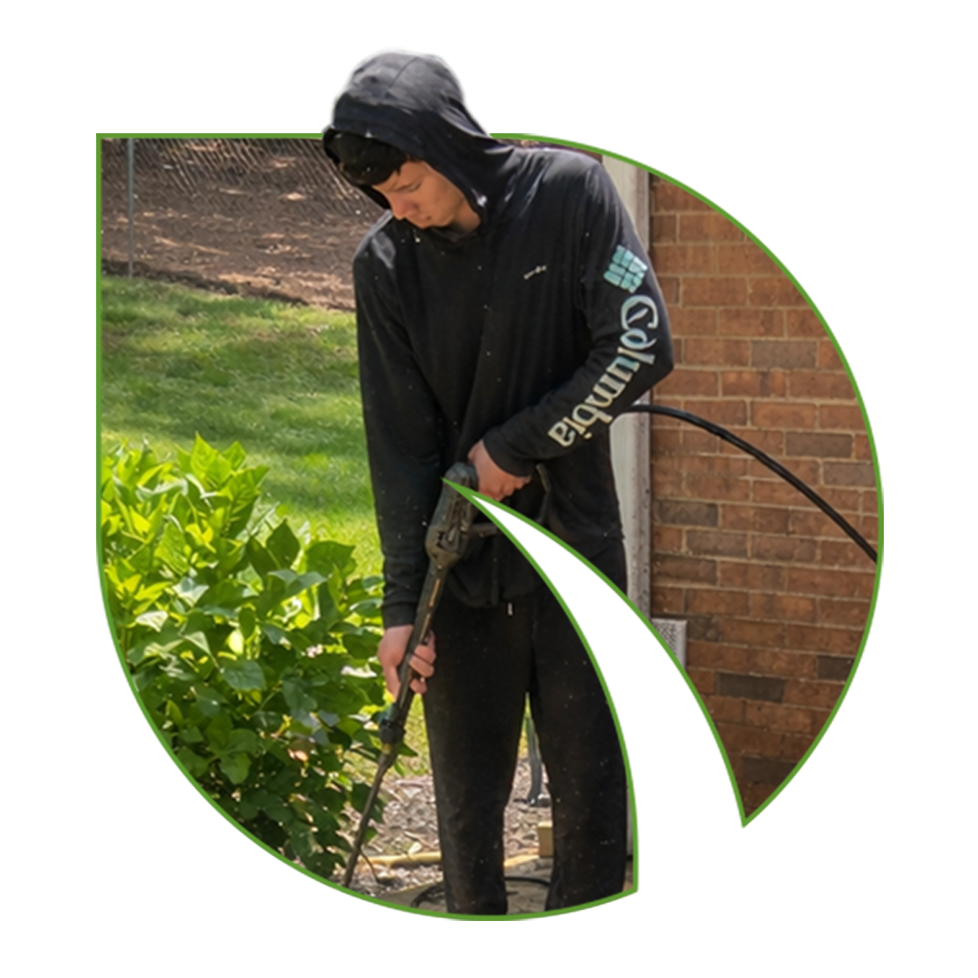 Joven trabajando en jardinería, usando una manguera para regar plantas cerca de una pared de ladrillos.