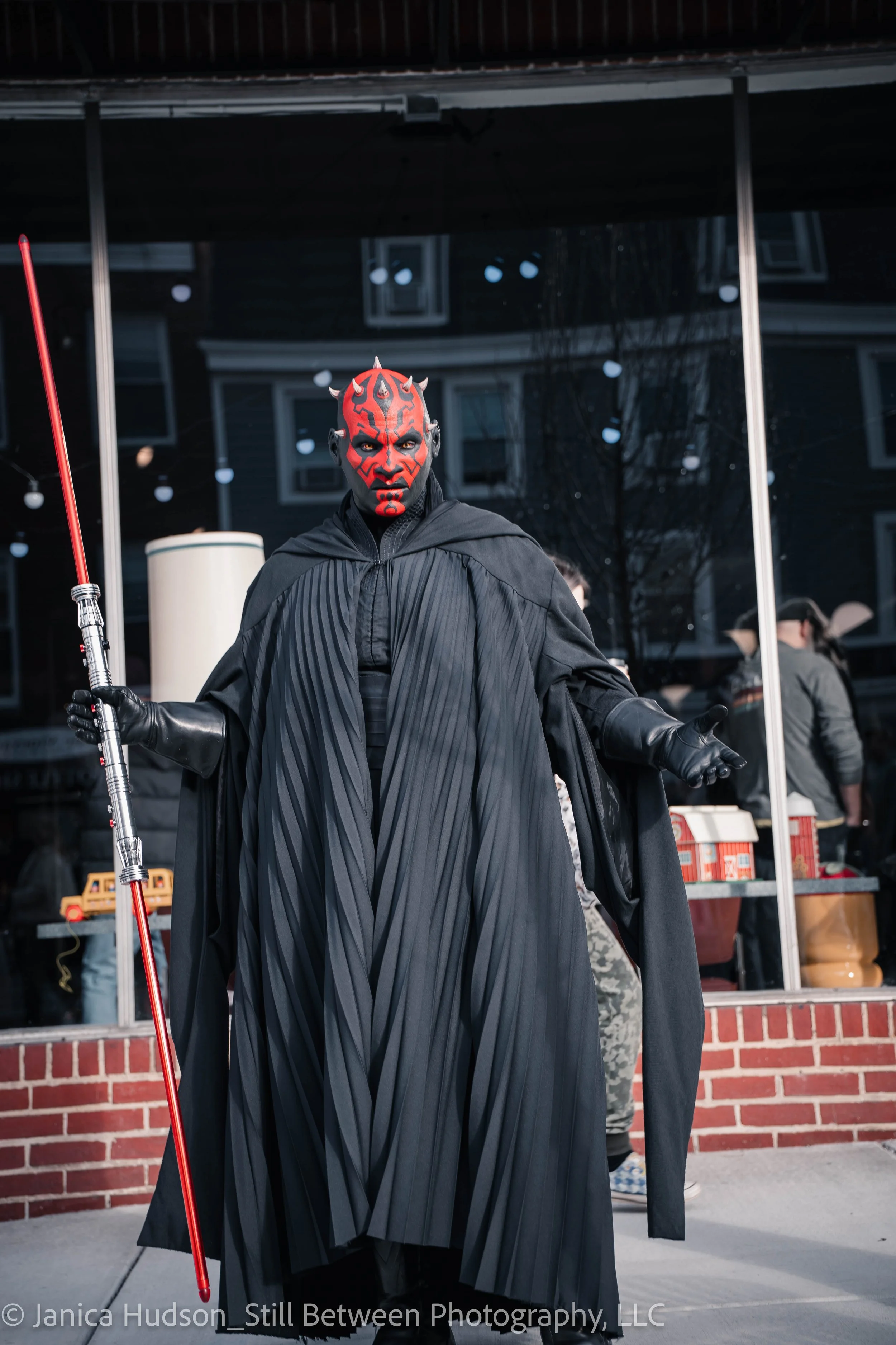 Person dressed as Darth Maul from Star Wars, wearing red and black face paint, standing outdoors in front of a van. The individual holds a red staff and wears a black cloak with pleated details.