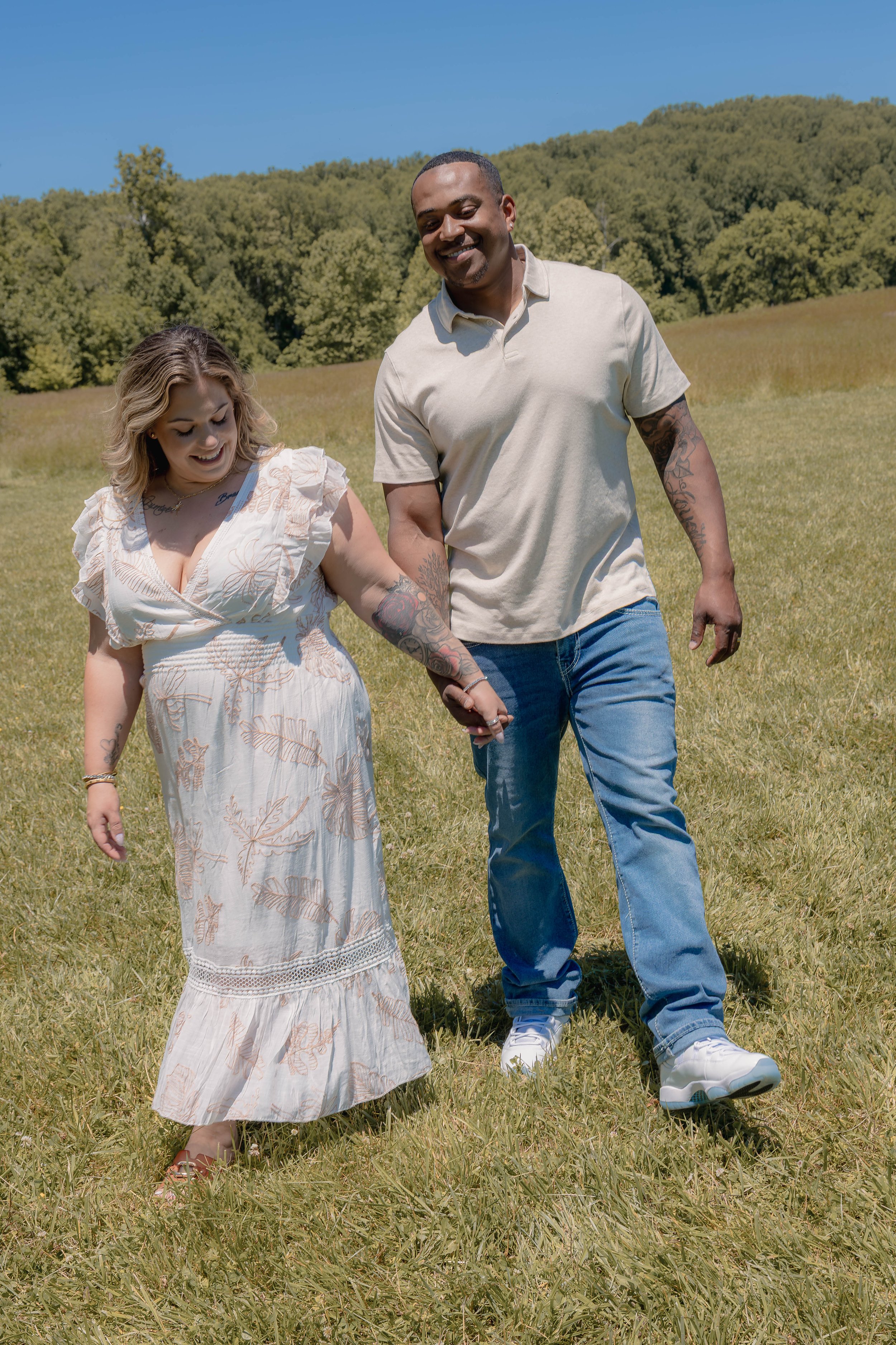 A couple walking outdoors in a grassy field on a sunny day, holding hands and smiling.