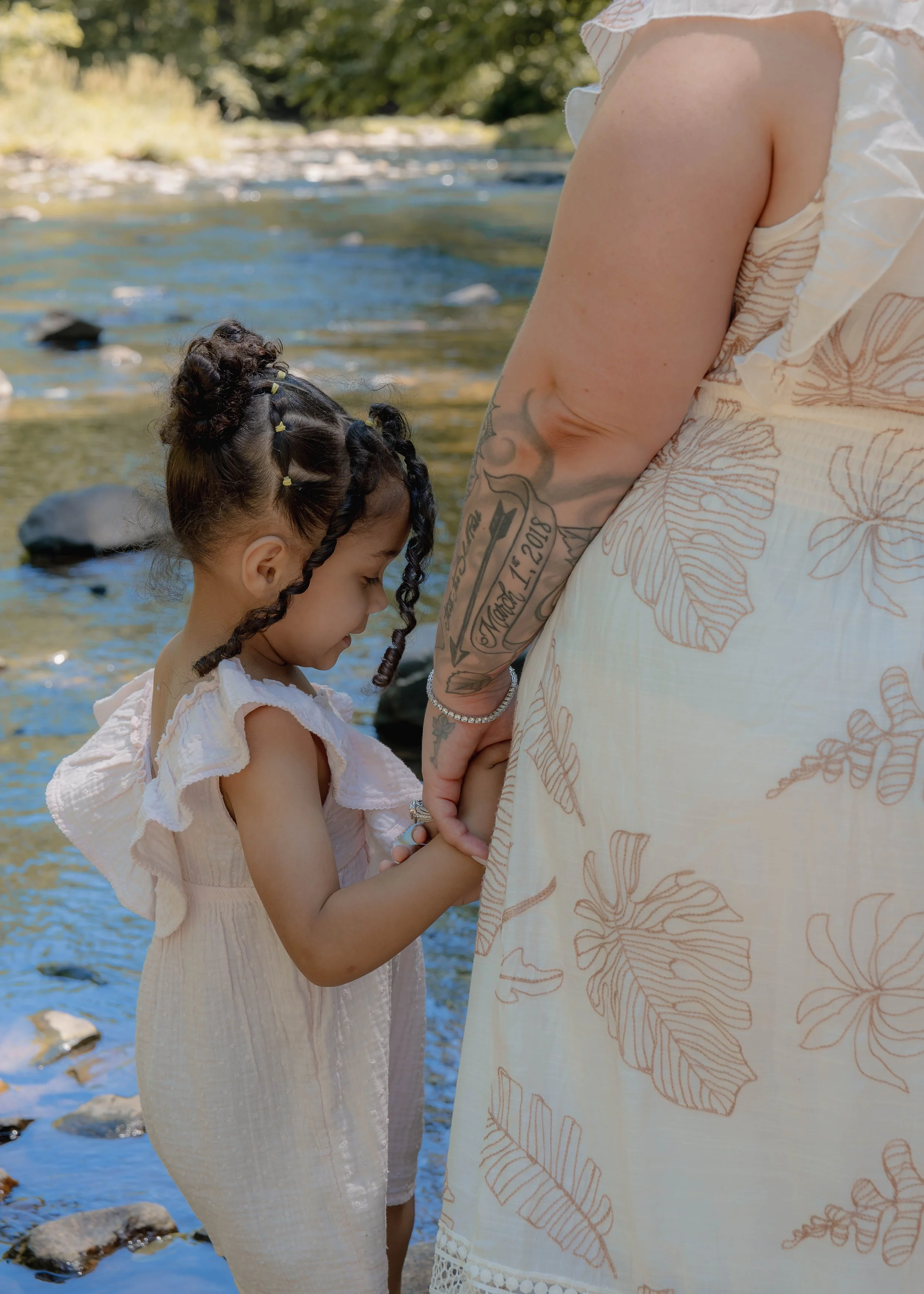 A young girl with braids and a white dress stands with a woman by a rocky riverbank, holding hands in a tender moment.