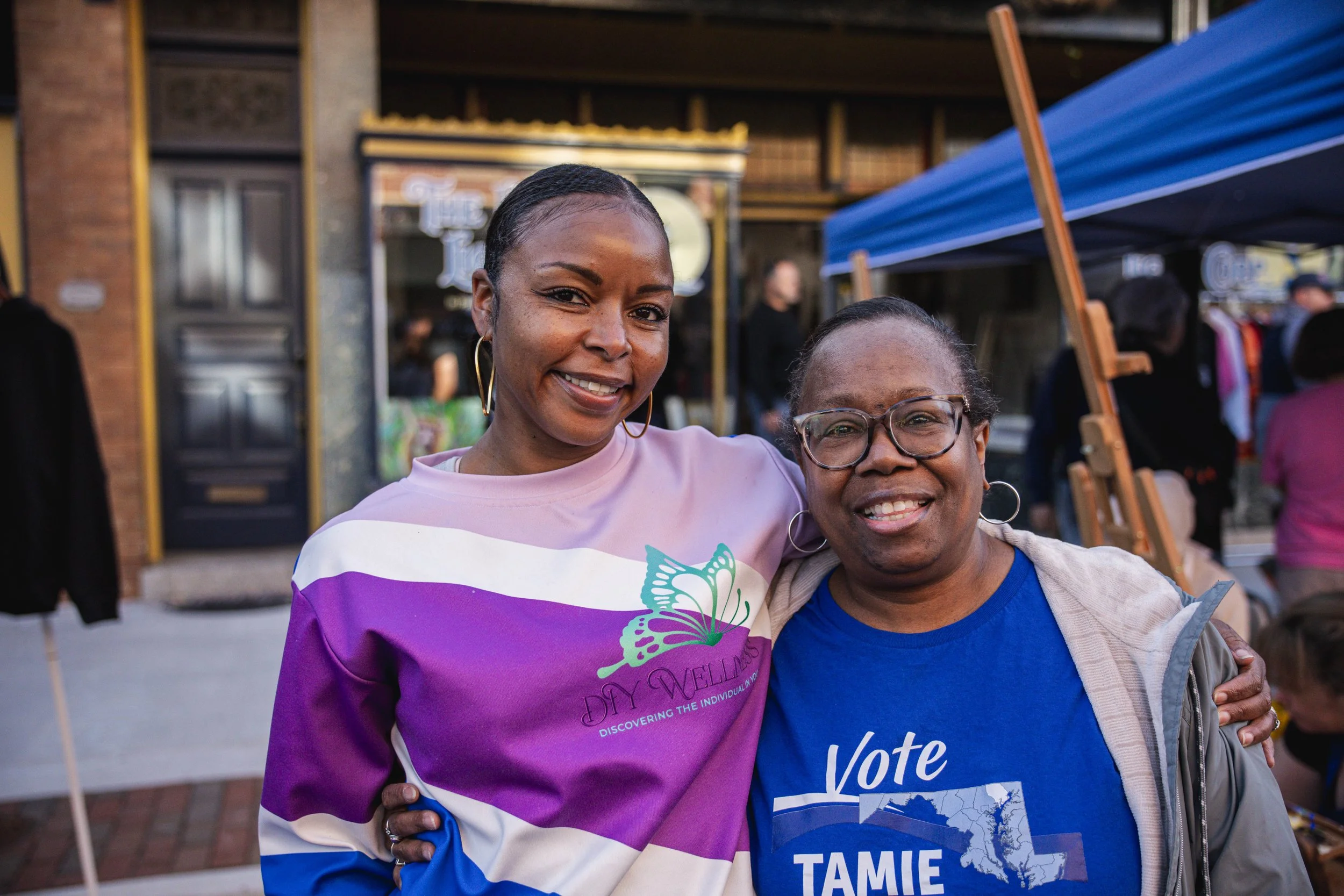 Two women smiling at an outdoor event, one wearing a pink, purple, and white shirt, the other in a blue shirt with 'vote' printed on it, people and booths in the background.