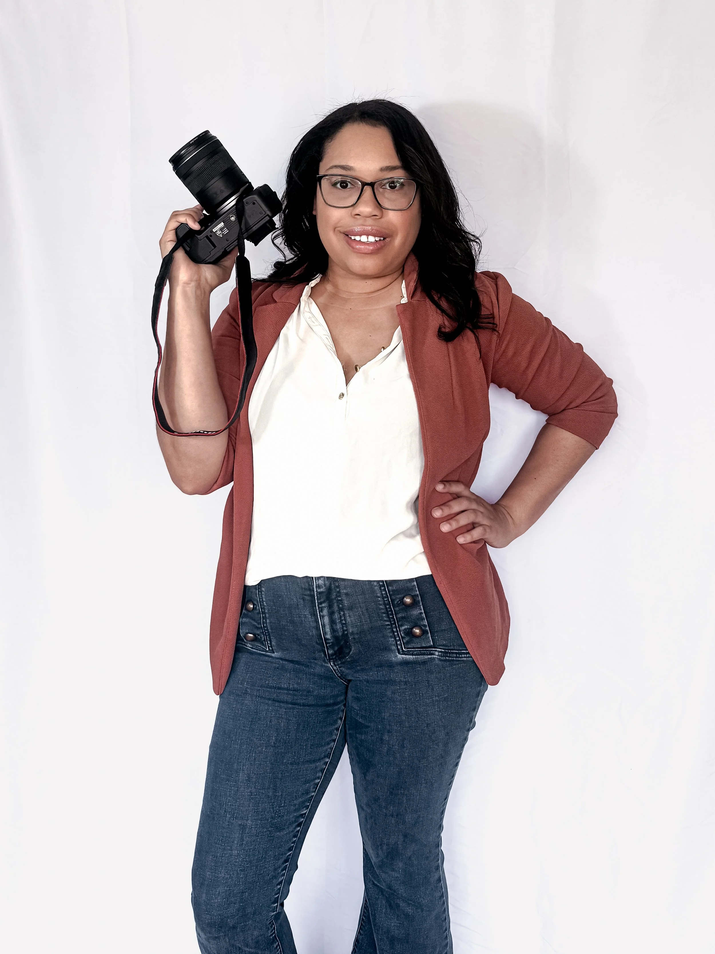 Woman with glasses and dark hair holding a camera on her shoulder, standing against a white background.