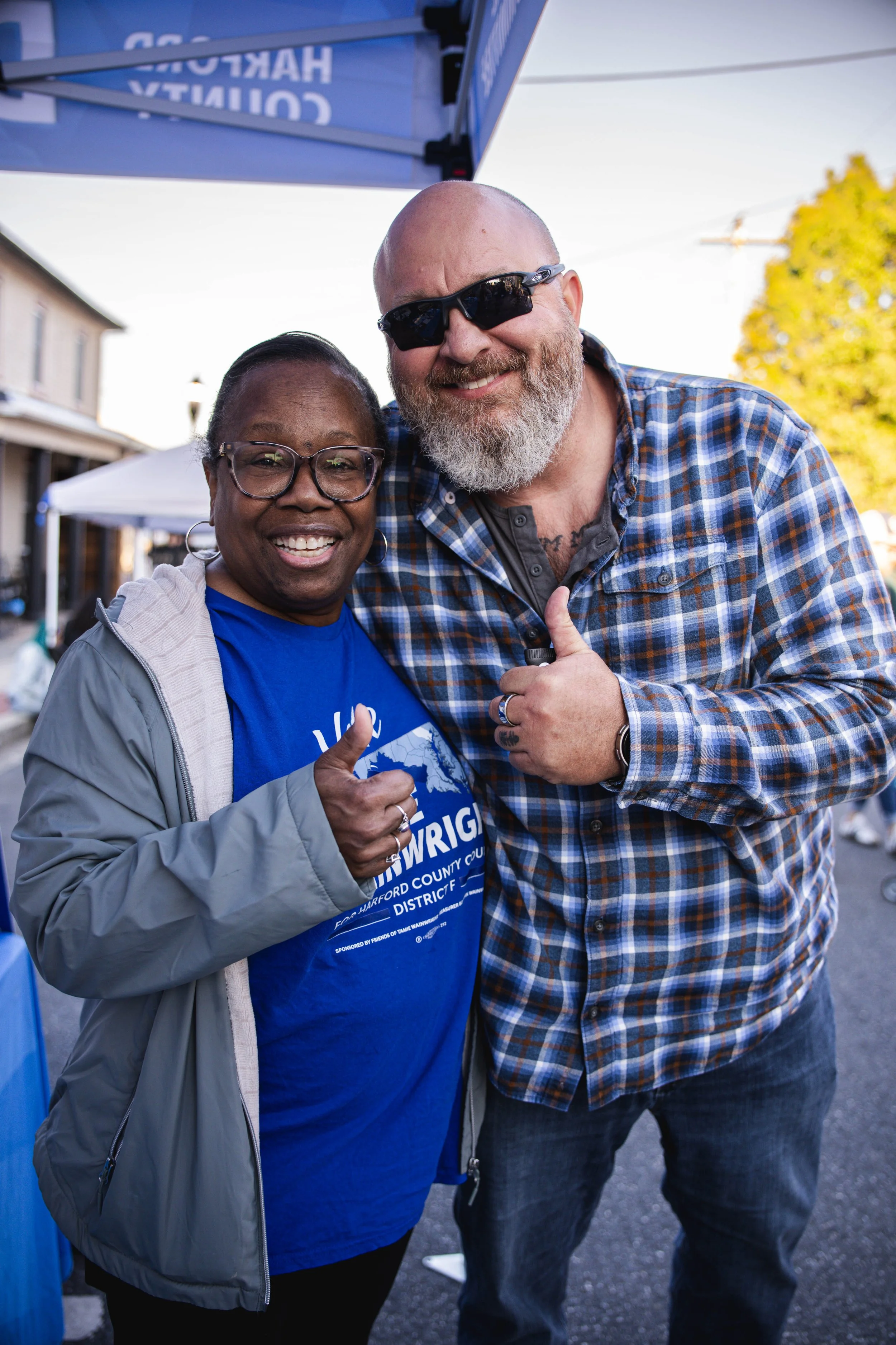 Two smiling people, a woman and a man, posing together outdoors at a community event. The woman is wearing glasses and a gray jacket, and the man is wearing sunglasses and a blue plaid shirt. They are giving thumbs-up gestures and standing under a bl