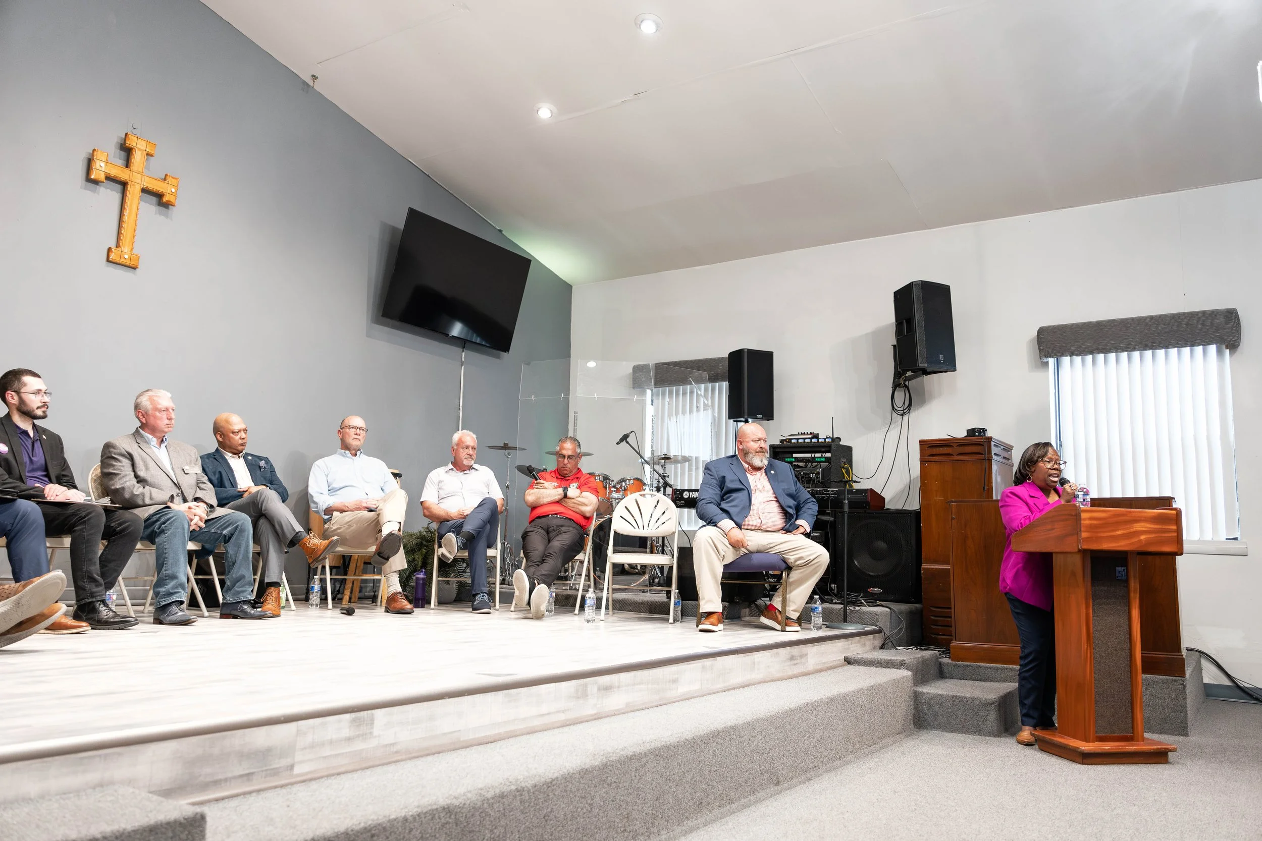 A group of nine diverse people seated on chairs on a stage, listening to a woman speaking at a wooden podium. The stage has a gray wall with a wooden cross and a large flat-screen TV mounted on it. There are musical instruments and speakers in the ba