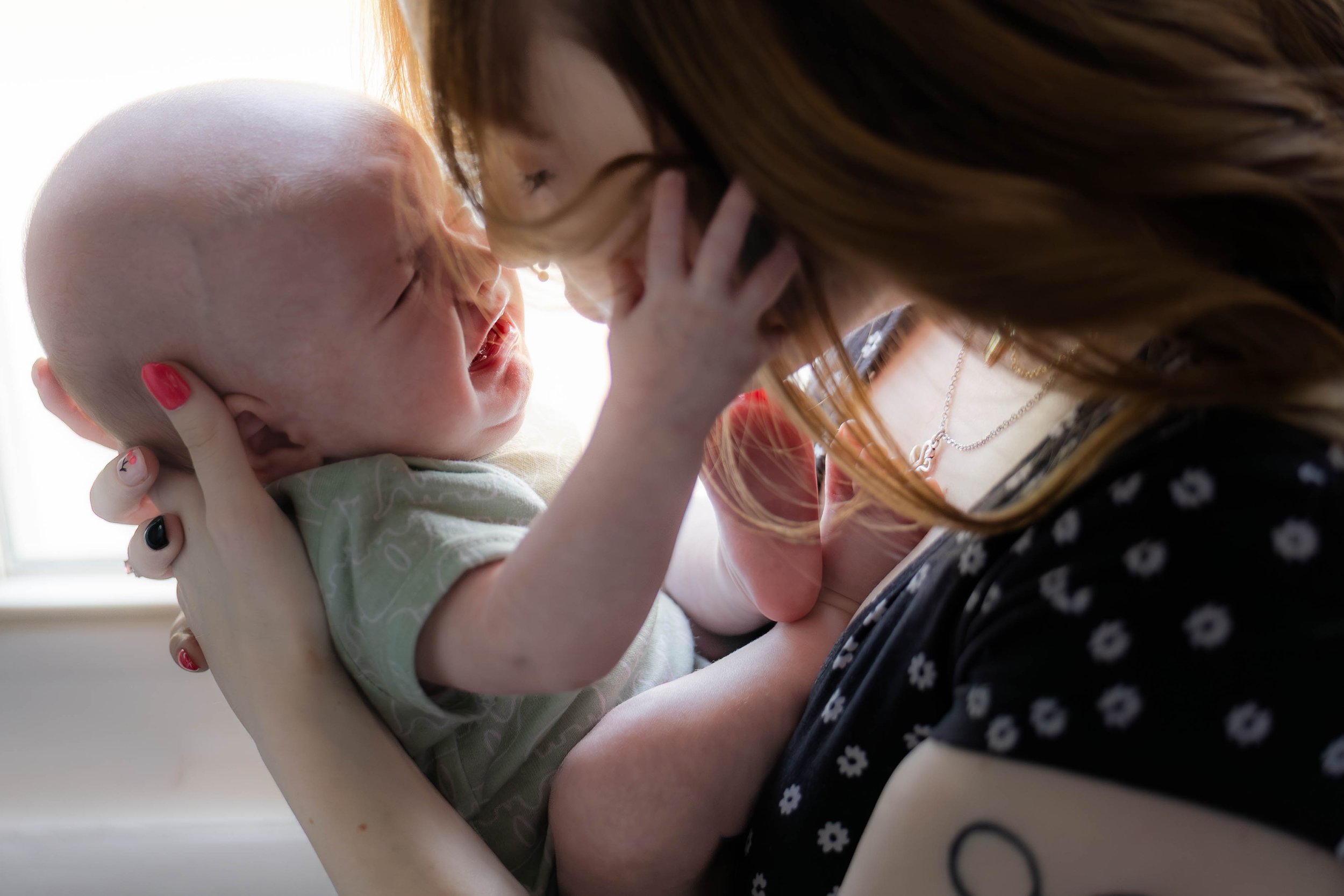 A woman holding a crying baby close to her face, in an indoor setting with a bright window light in the background.