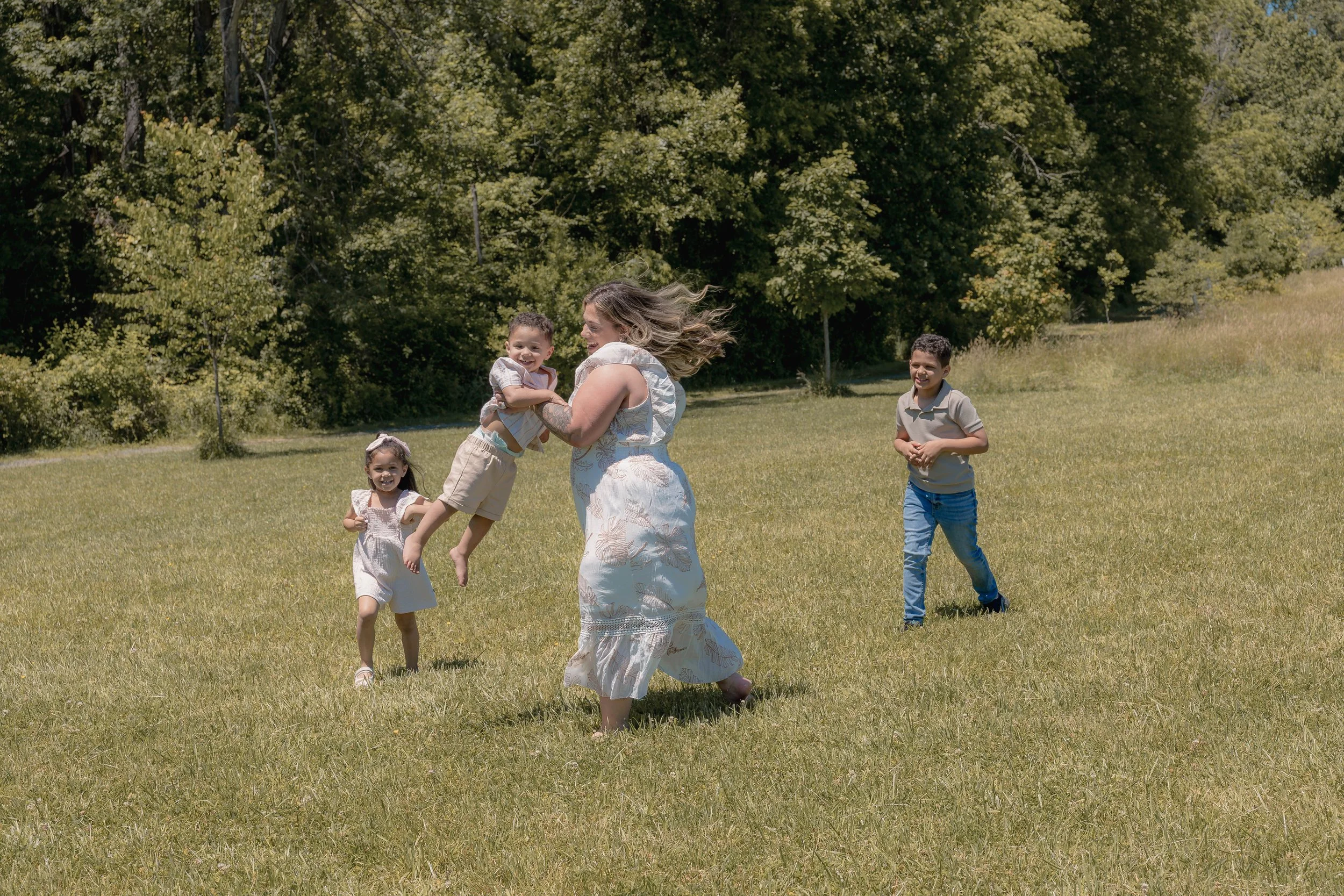 A woman and three children playing and running on a grassy field with trees in the background on a sunny day.