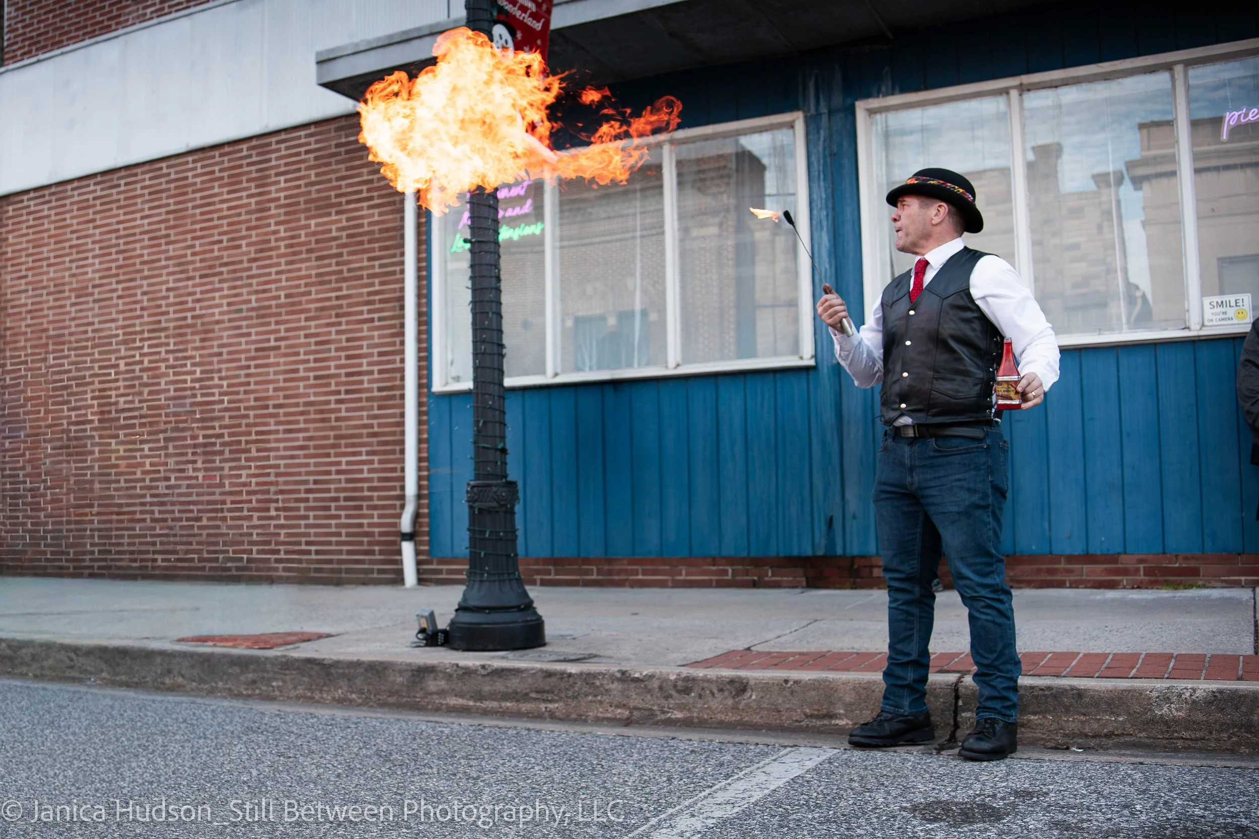 A man dressed in vintage attire, including a black hat, white shirt, red tie, and black vest, is blowing fire from his mouth as part of a street performance. He holds a matchstick in one hand and a bottle of fire-eating liquid in the other. The backd