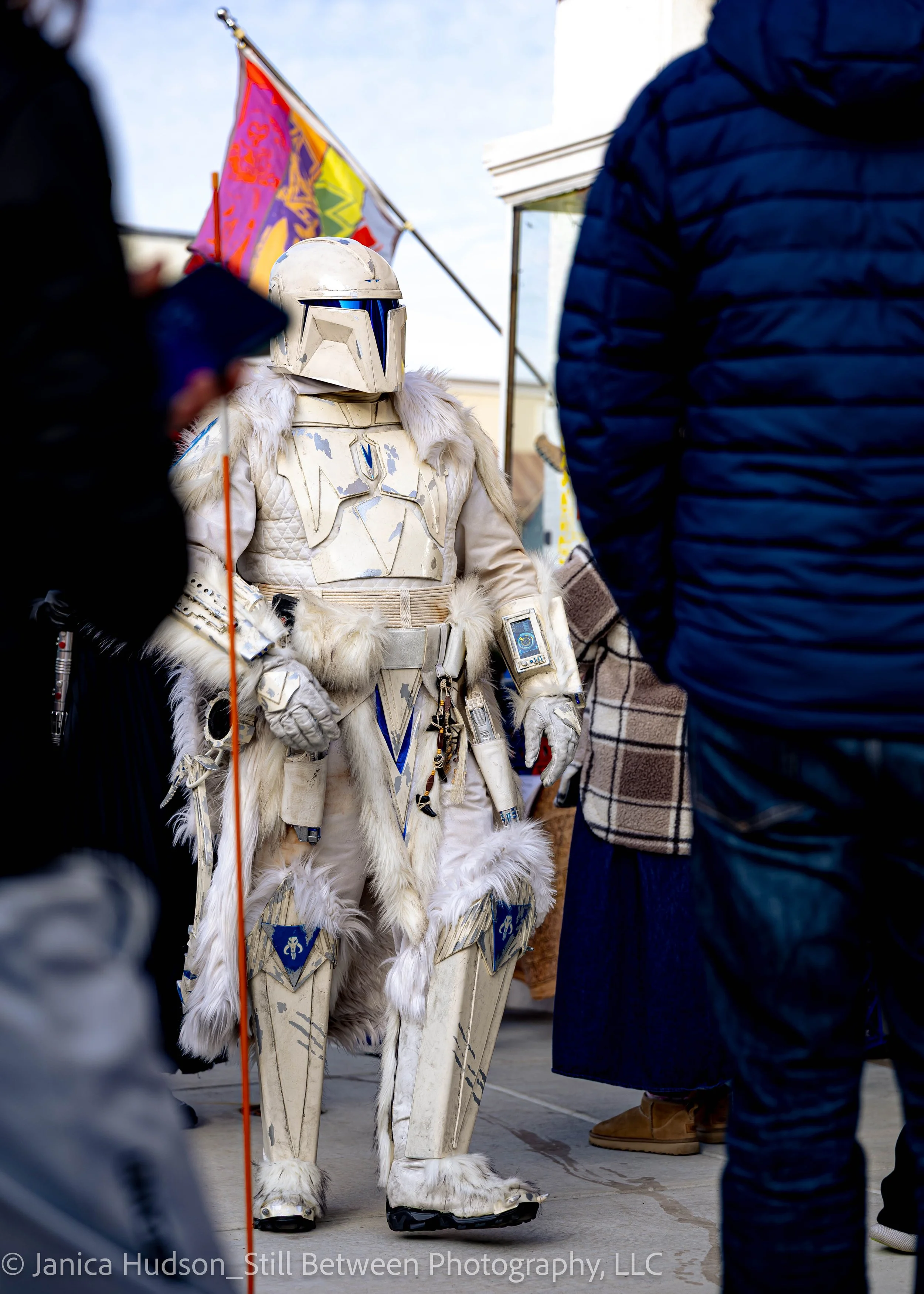 A person in a Star Wars scout trooper costume standing among people at an outdoor event, with a purple and yellow flag in the background.
