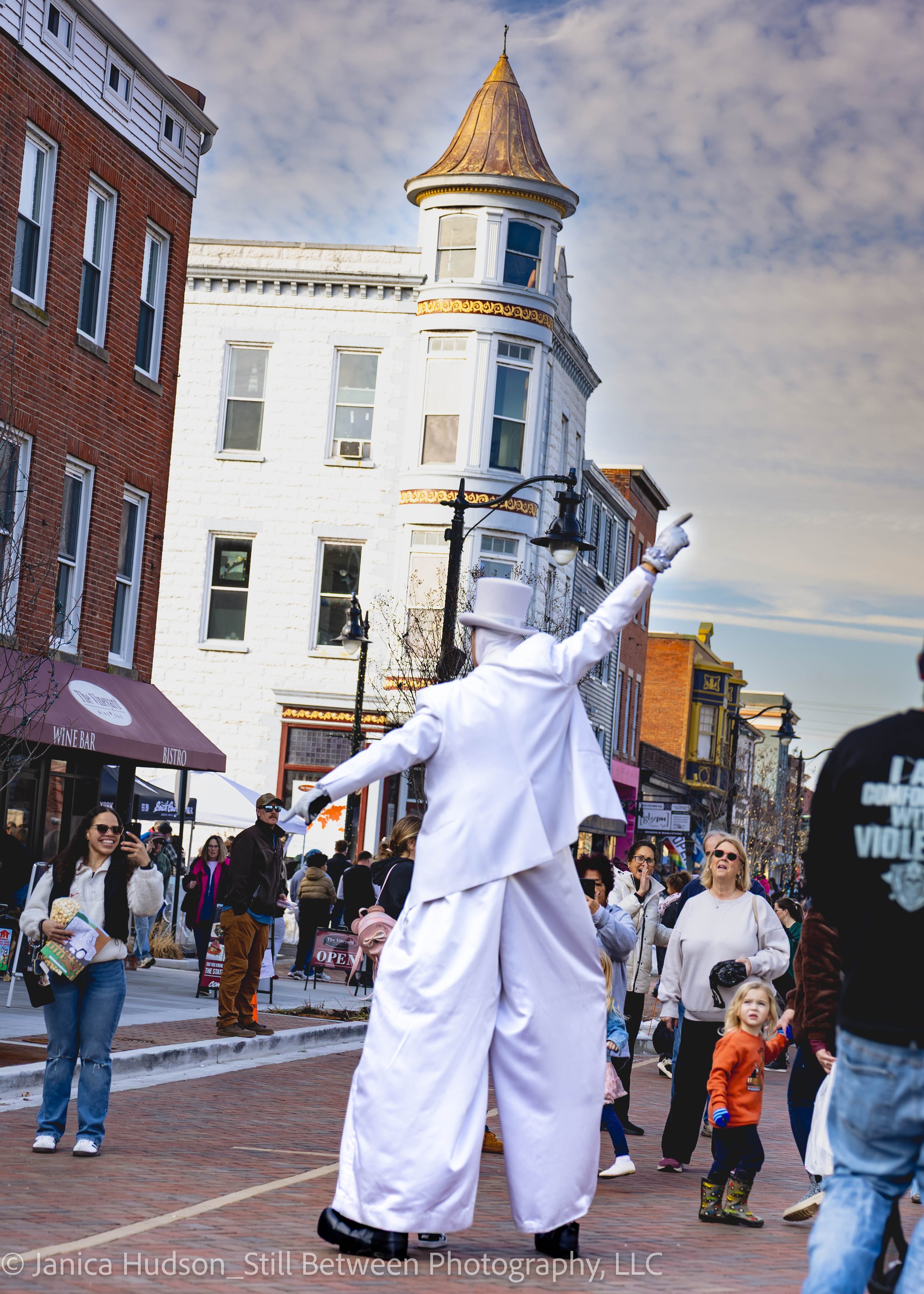 Street scene during a festival with a person dressed as a mime on stilts in a white costume, with a crowd of spectators and participants, multi-story buildings, and a cloudy sky.