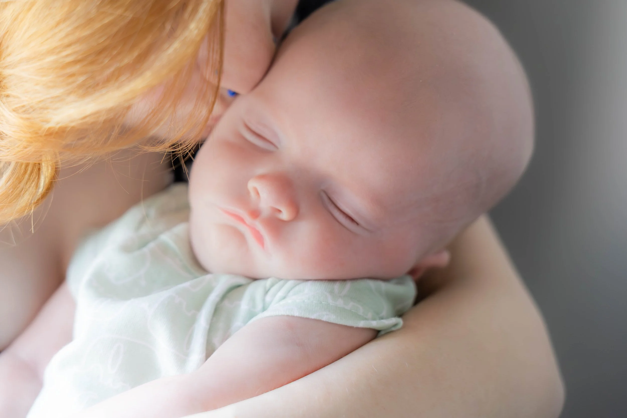Close-up of a baby sleeping in an adult's arms, with an adult with red hair and a light green patterned shirt holding the baby.