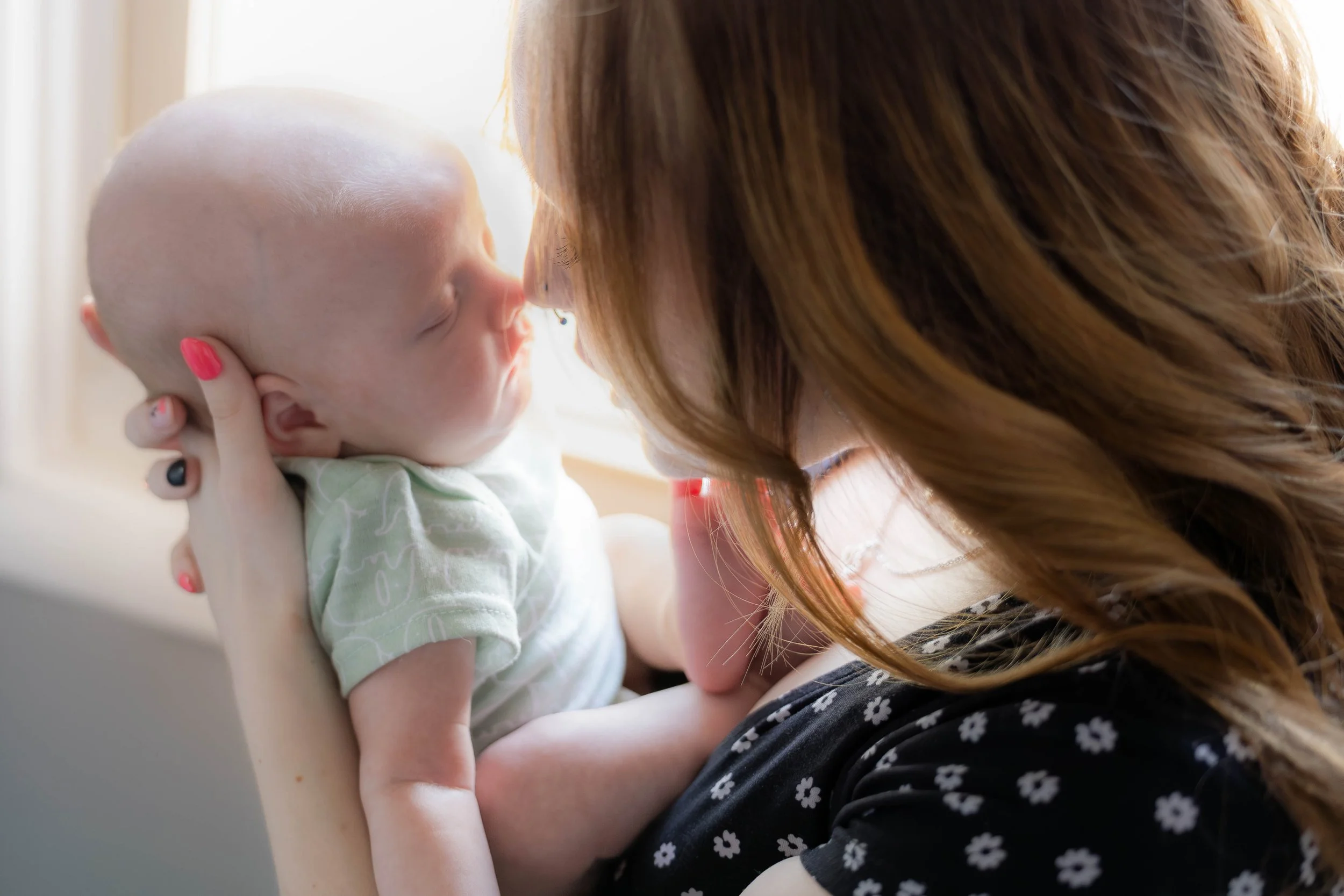 A woman with long hair holding a baby close, touching their faces gently, with light streaming in from a window.