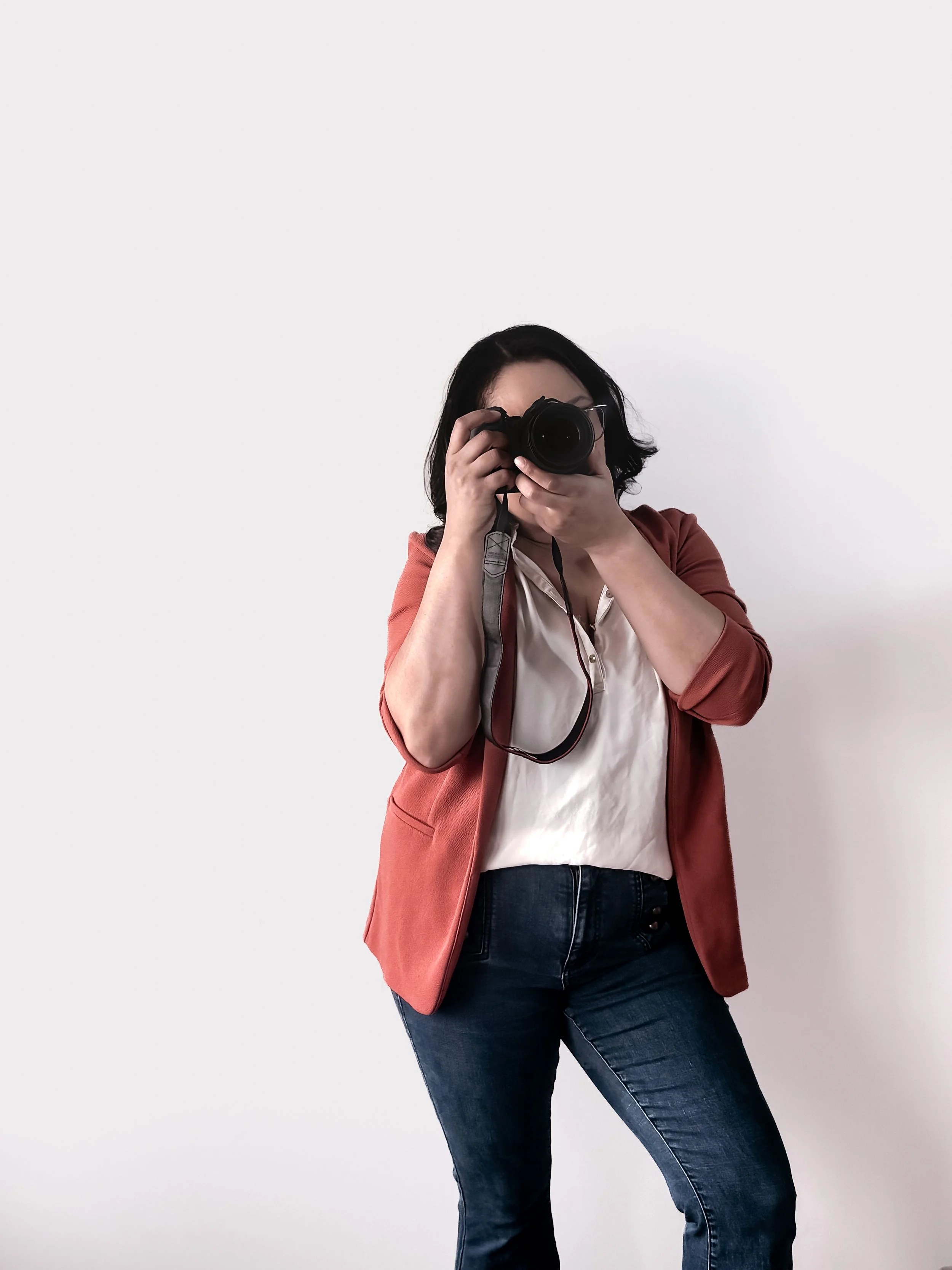 Woman with dark hair taking a photo with a camera against a plain white wall, wearing a reddish-brown blazer, white blouse, and dark jeans.