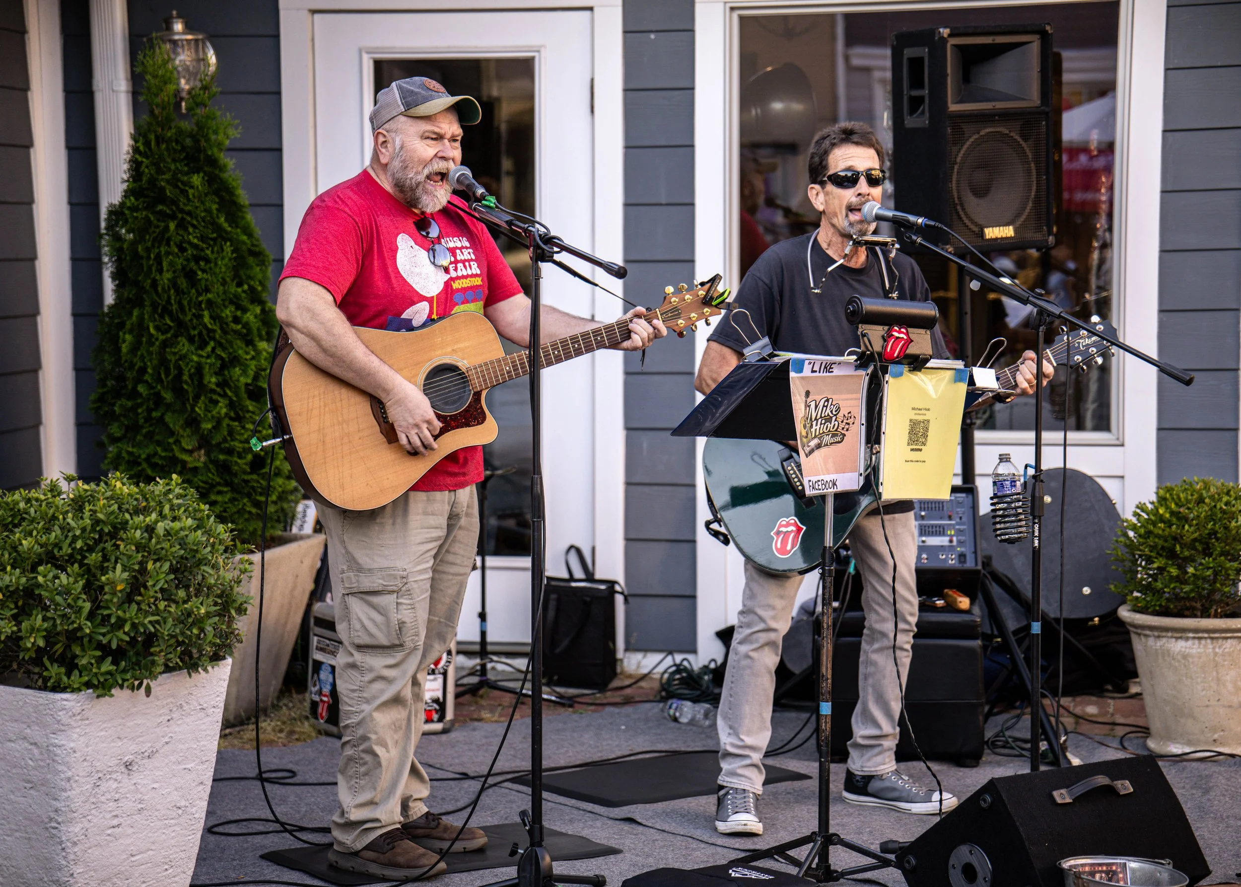 Two men performing on an outdoor stage with microphones and guitars, one singing and the other playing guitar, in front of a gray house with potted plants, sound equipment, and a Yamaha speaker.