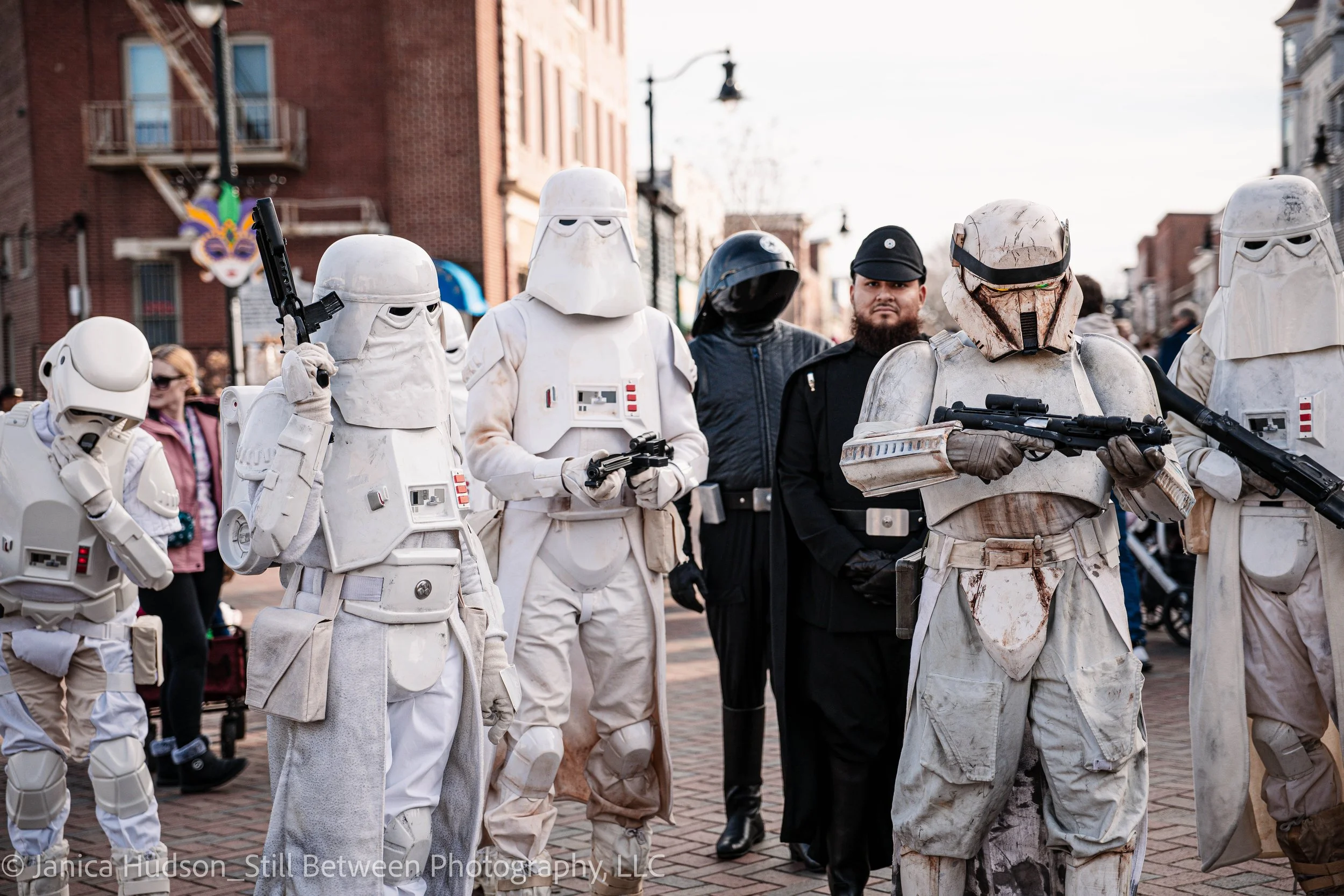 A group of people dressed as Star Wars stormtroopers standing on a street, with some holding blasters, and a man dressed as a police officer standing among them. People and buildings are visible in the background.