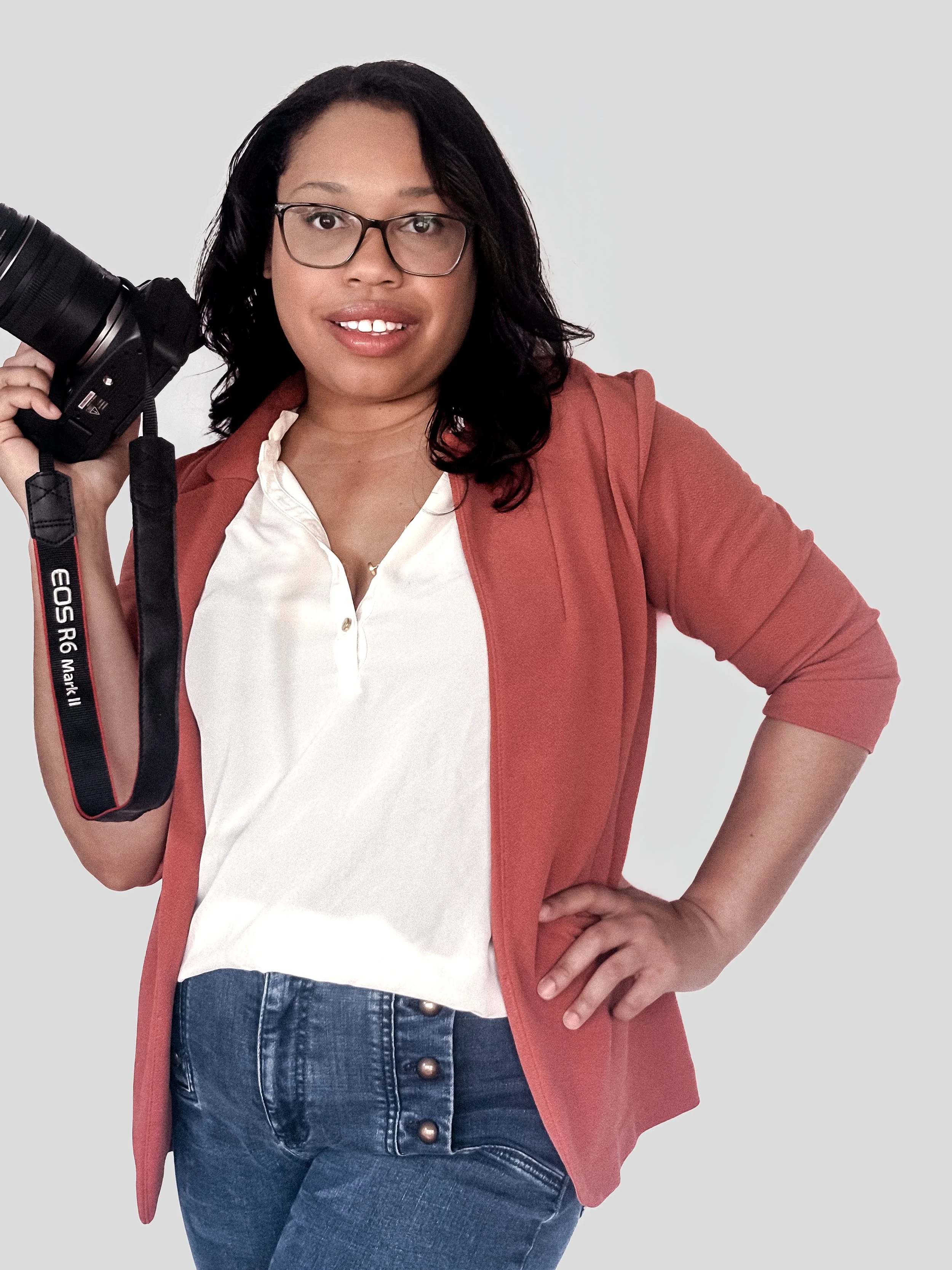 A woman with glasses and long dark hair holding a camera on her shoulder, standing with her hand on her hip against a plain background.
