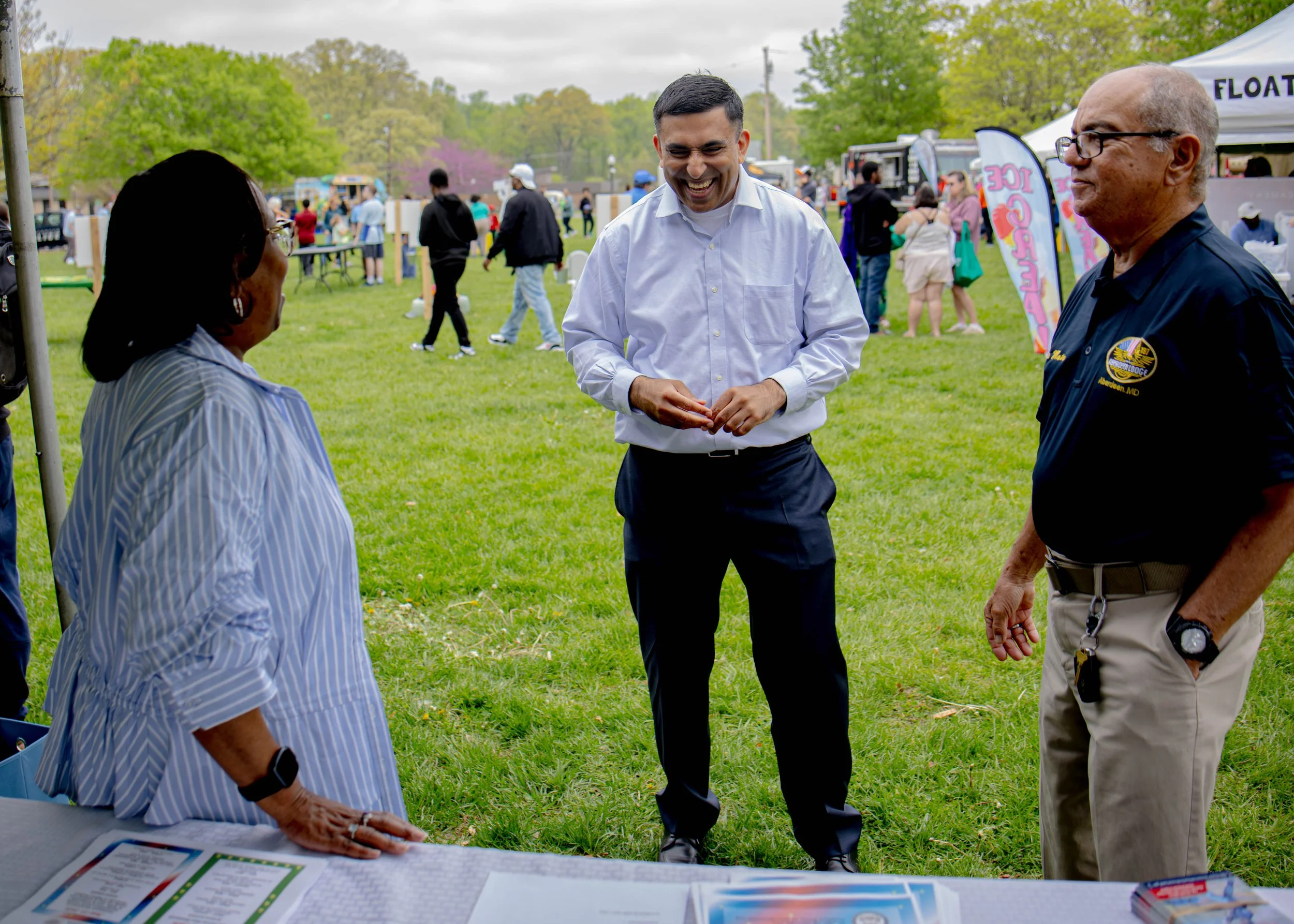 People at an outdoor event talking and smiling, with food and informational tables and tents in the background.