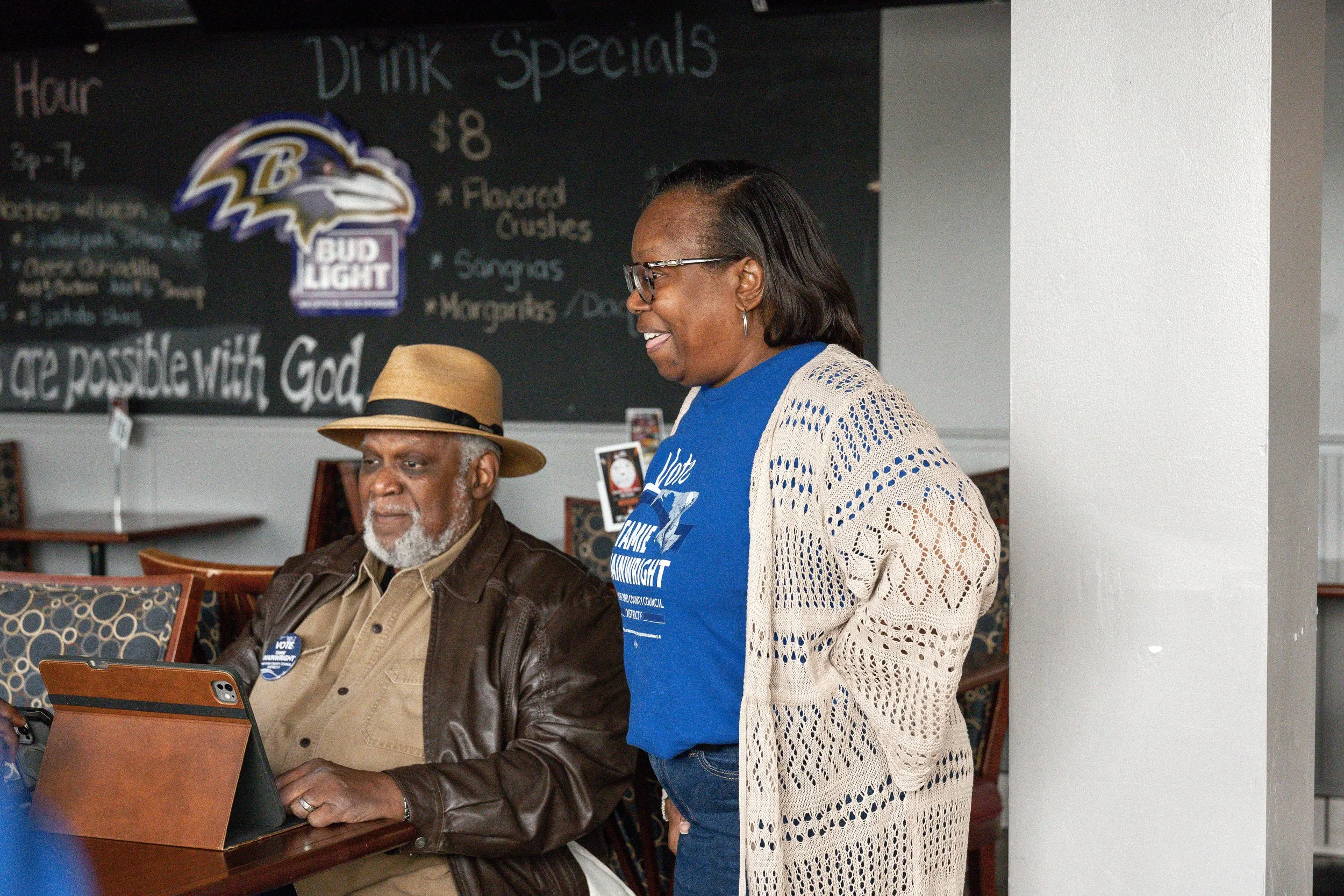 A woman with glasses and a beige crochet cardigan smiling while talking to an elderly man with a gray beard wearing a hat and brown leather jacket. They are in a cafe or restaurant with a chalkboard menu and a Baltimore Ravens logo on the wall.
