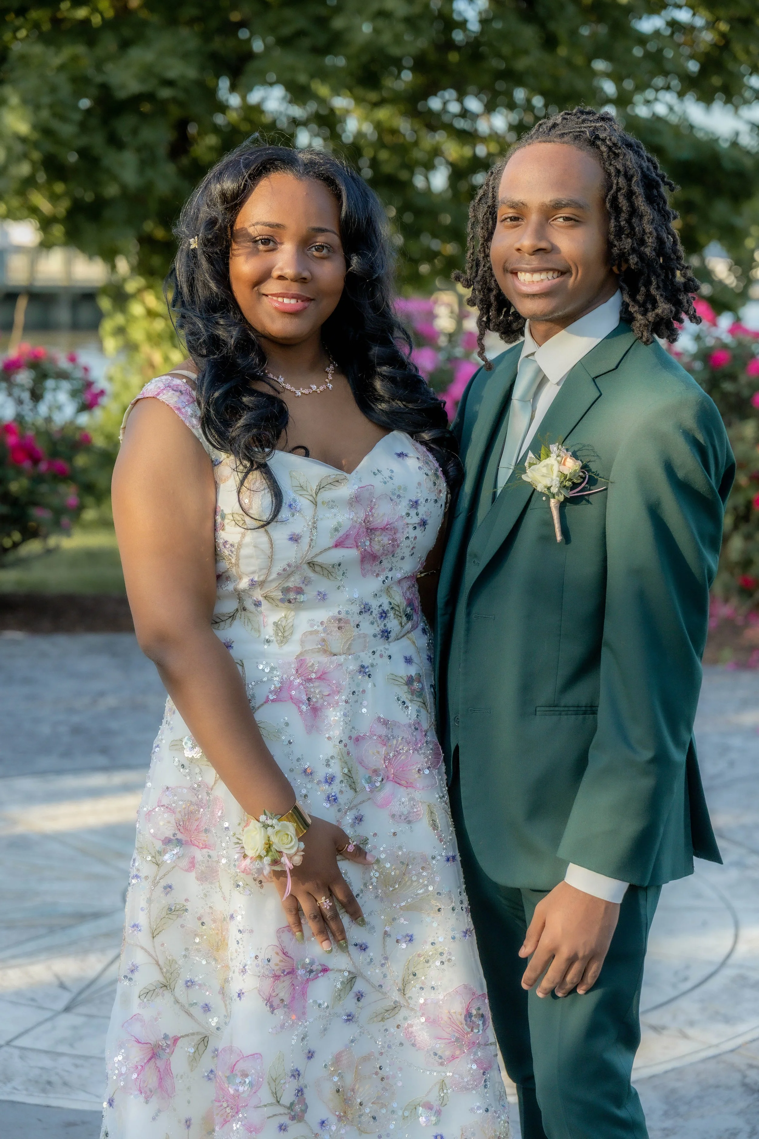 A couple dressed in formal wedding attire standing outdoors with trees and pink flowers in the background, smiling at the camera.