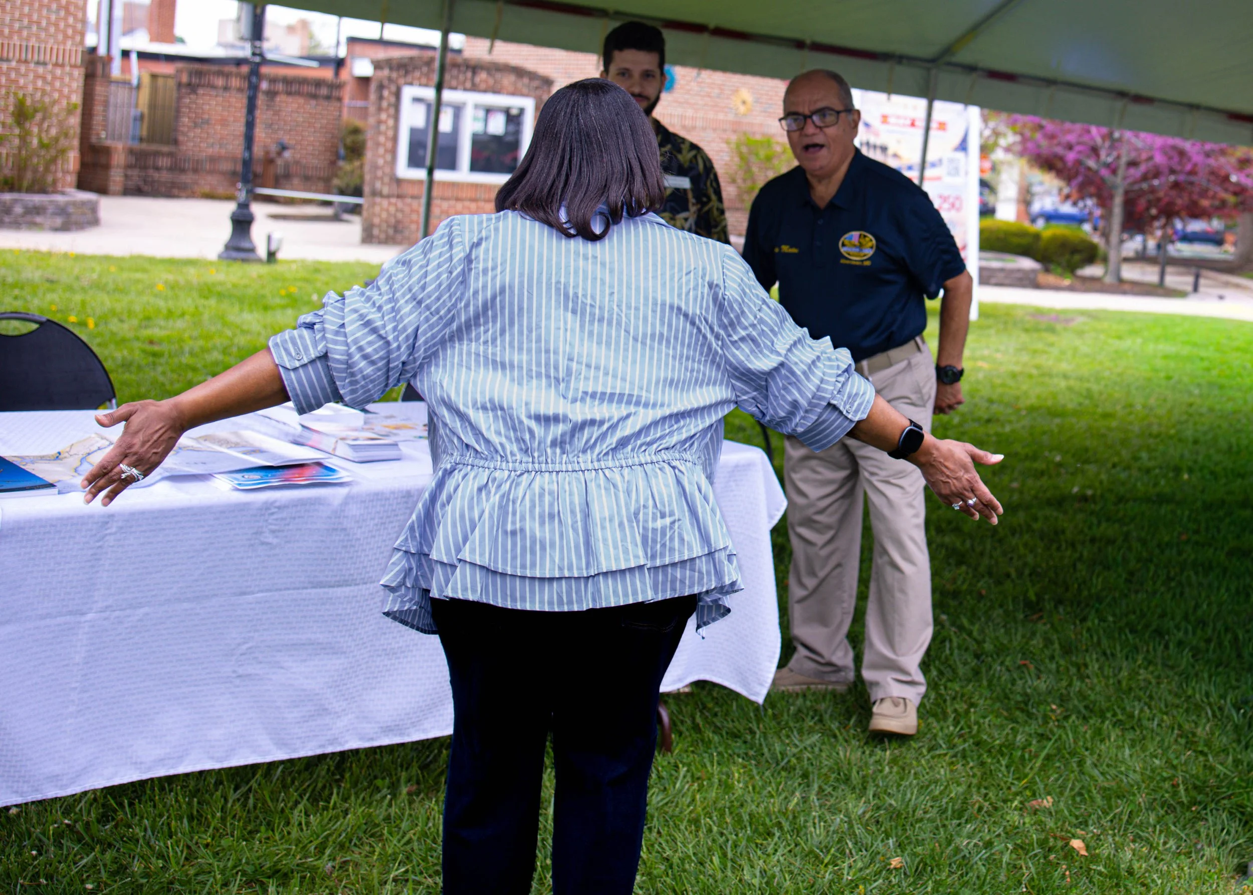 A woman with shoulder-length dark hair wearing a blue and white striped shirt with arms outstretched, standing at an outdoor event table. Behind her, two men are engaged in conversation under a green canopy, with one man wearing glasses and a navy sh