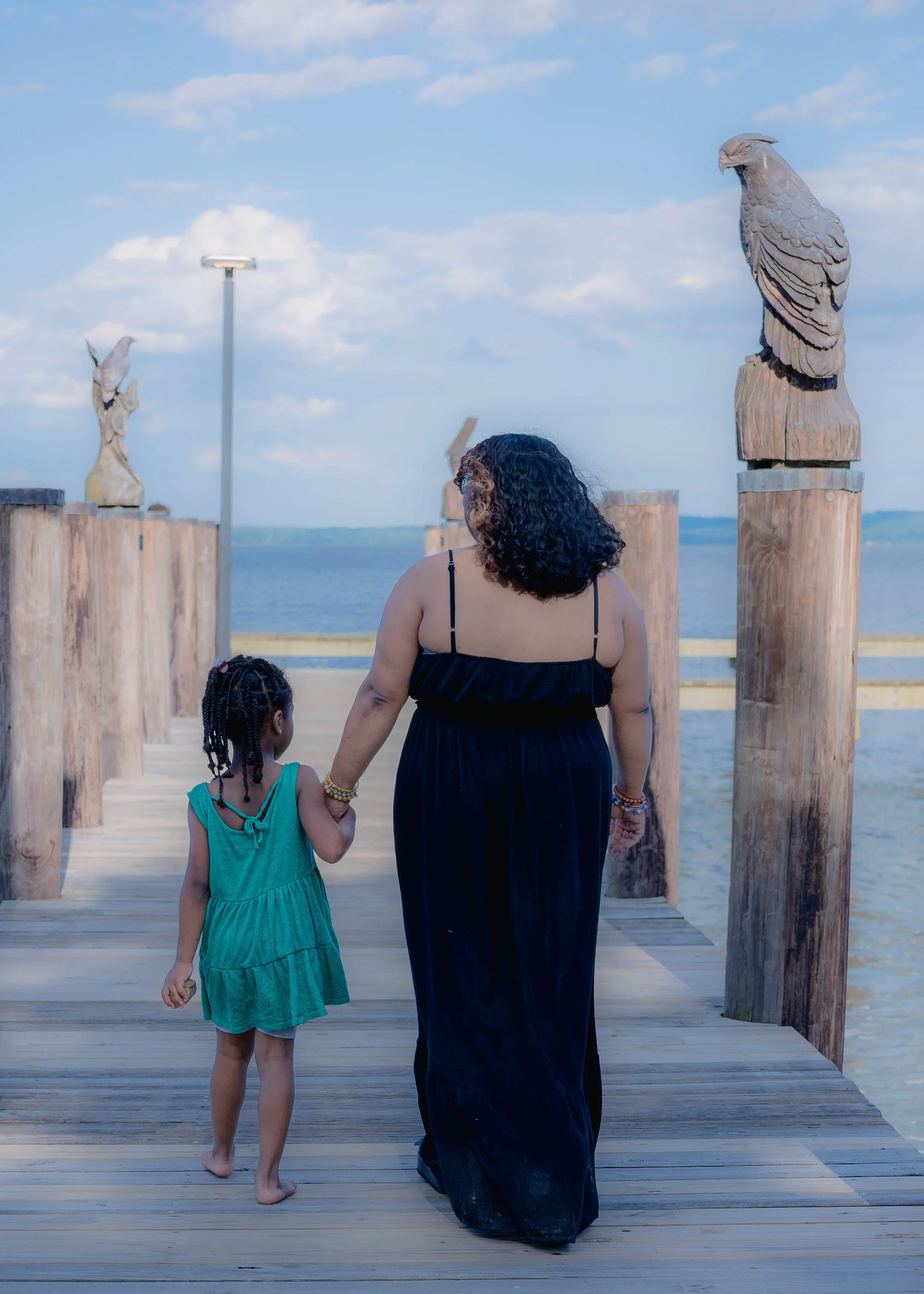 A woman and a girl walking on a wooden pier towards the water, with wooden eagle sculptures on posts along the pier, under a partly cloudy sky.
