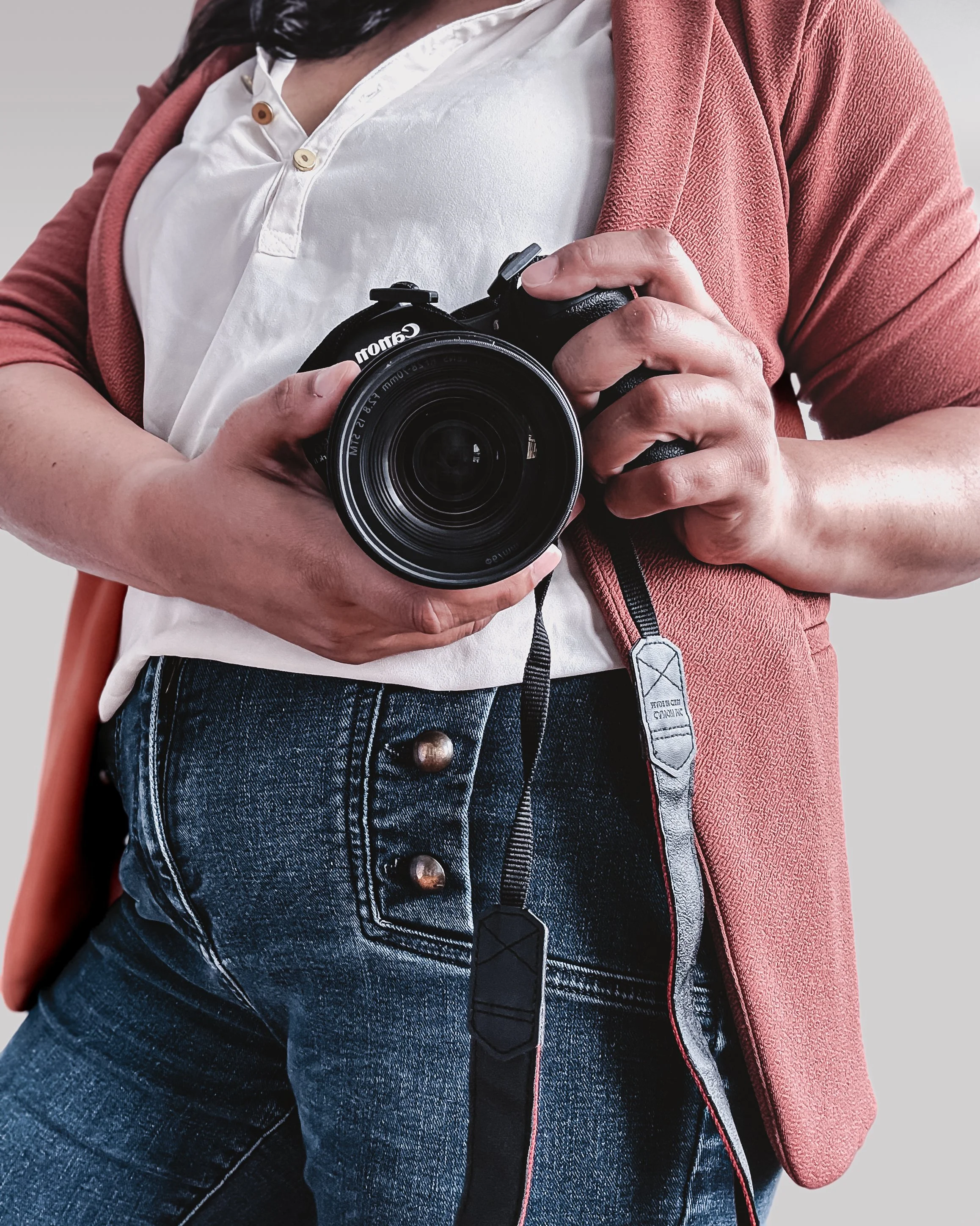 Person holding a Canon camera, taking a photo, wearing a white shirt, pink jacket, and jeans.