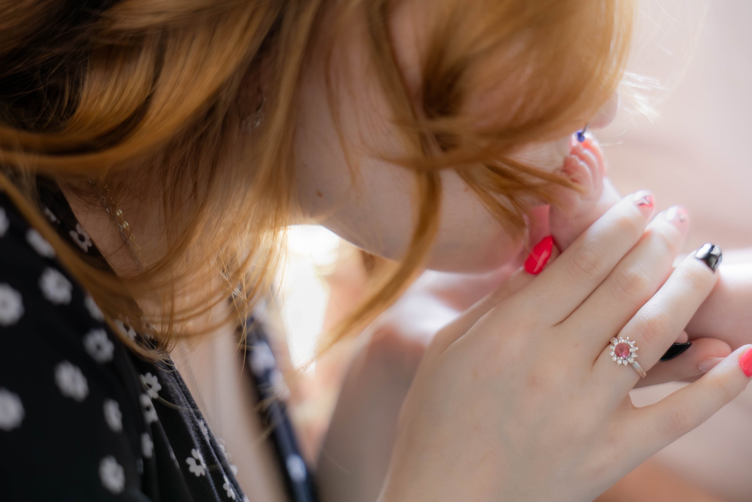 Close-up of a woman with red hair, wearing a ring with a pink gemstone surrounded by small diamonds, and holding her hands together near her face, appearing thoughtful or praying.
