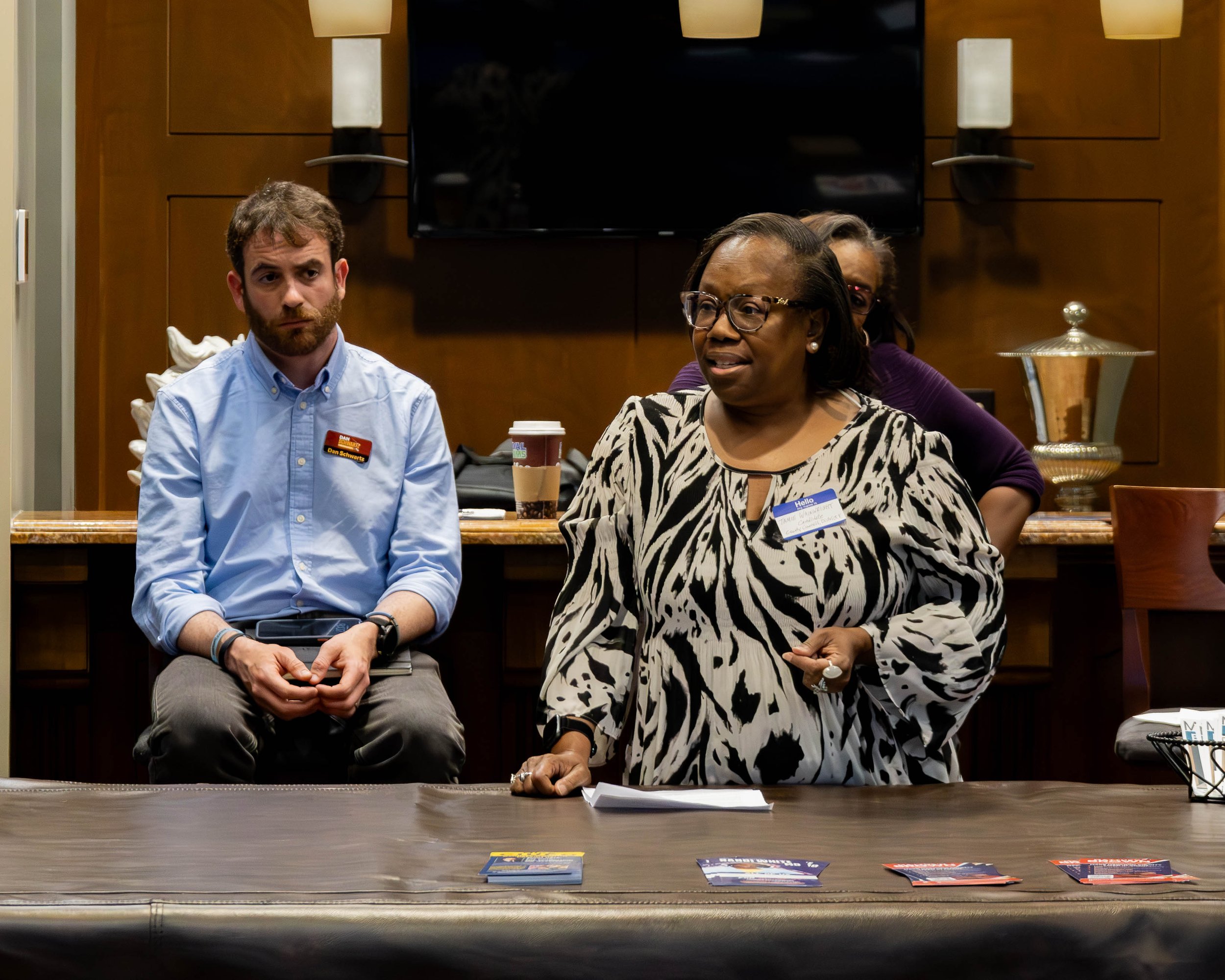 Three people seated at a table during a meeting, with pamphlets and papers spread out on the table in front of them. A man on the left with a beard, glasses, and a name tag reading "Dan Schwartz" wears a light blue button-up shirt. A woman in the mid