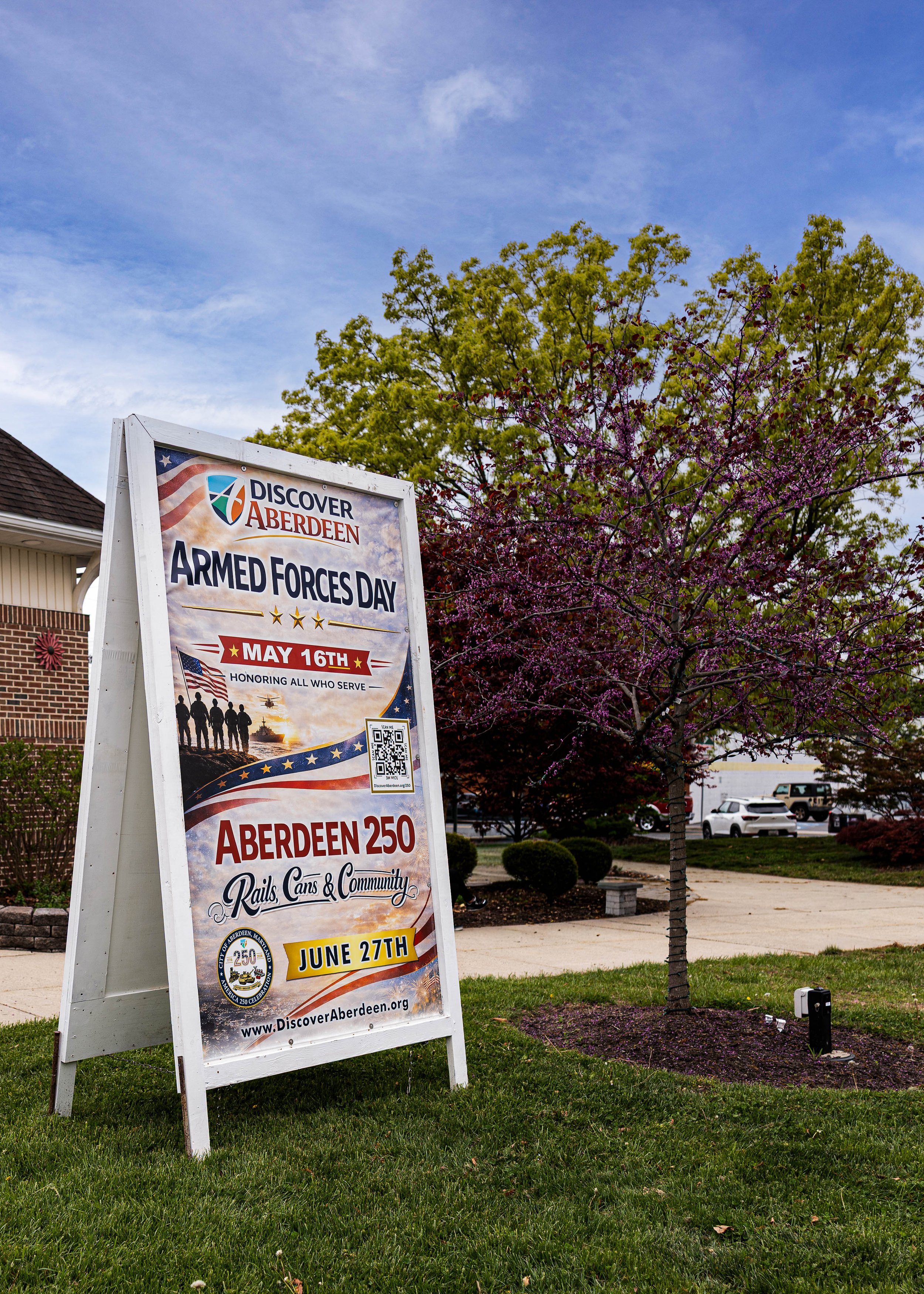 An outdoor scene with a sign promoting Armed Forces Day events in Aberdeen, Maryland, with trees and a parking lot in the background.