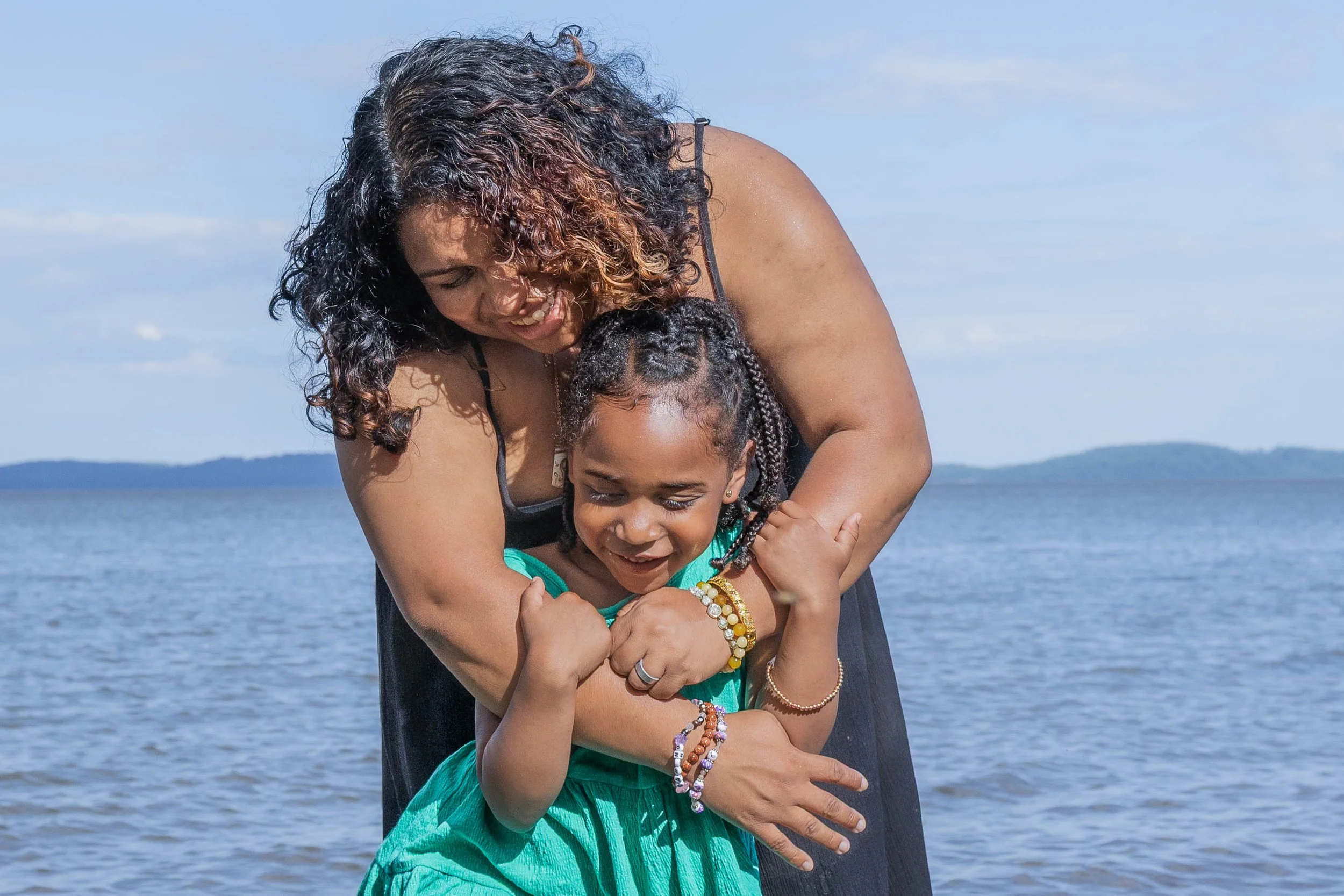 A woman with curly hair hugging a young girl with braided hair near a body of water with a distant shoreline and a blue sky.