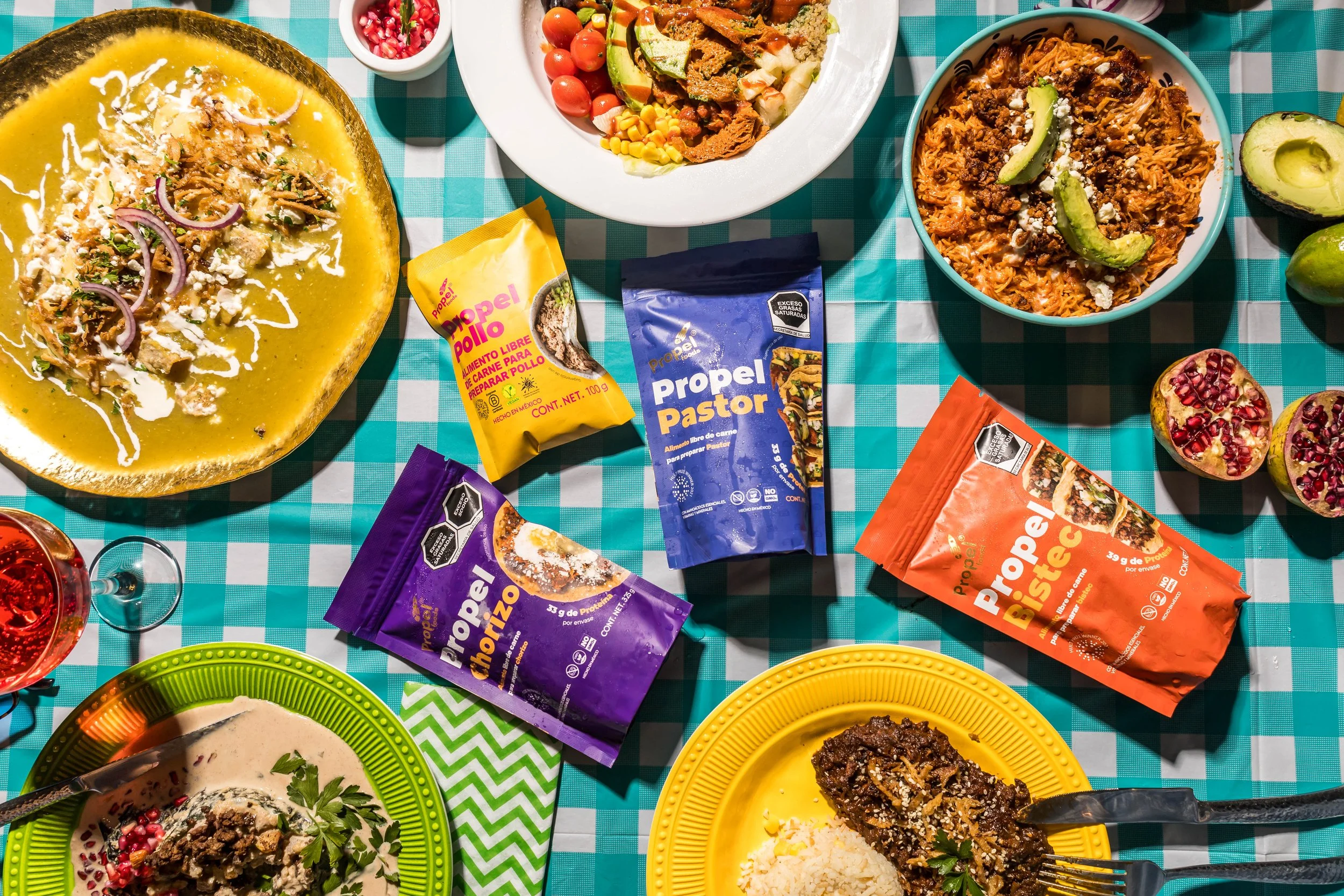 Mesa con varios platos de comida mexicana, incluyendo sopa, tacos, guacamole, arroz, y frutas, además de paquetes de productos Propel con diferentes sabores y un vaso de vino rosado.