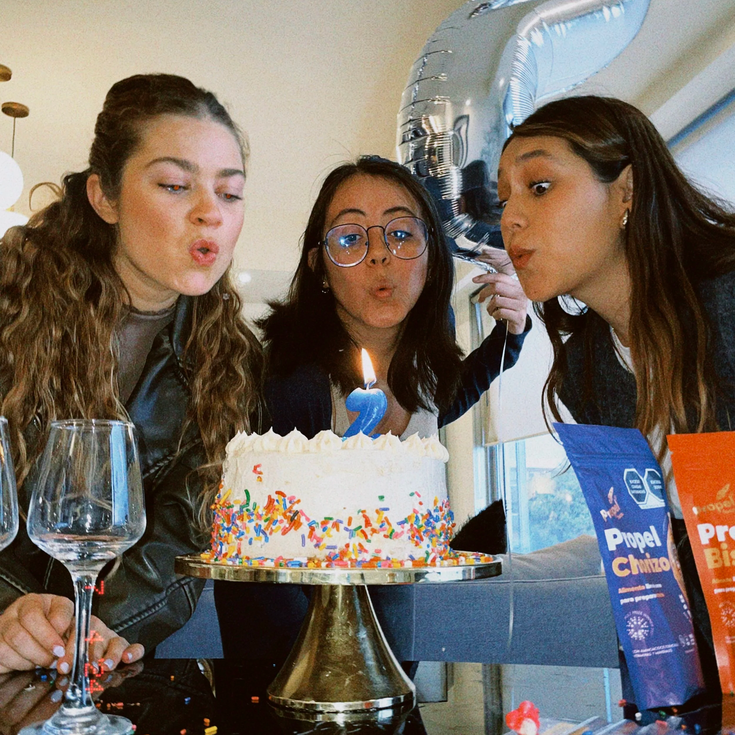 Tres mujeres celebrando con un pastel de cumpleaños decorado con glaseado blanco y confeti de colores, soplando las velas en forma de número 2, en un ambiente festivo con globos y bebidas en la mesa.