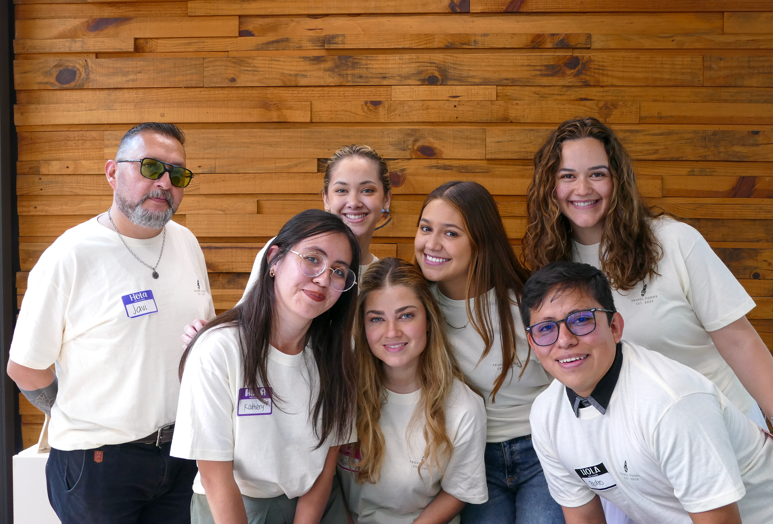 Grupo de siete personas sonriendo, varias con gafas, en una reunión con fondo de madera.