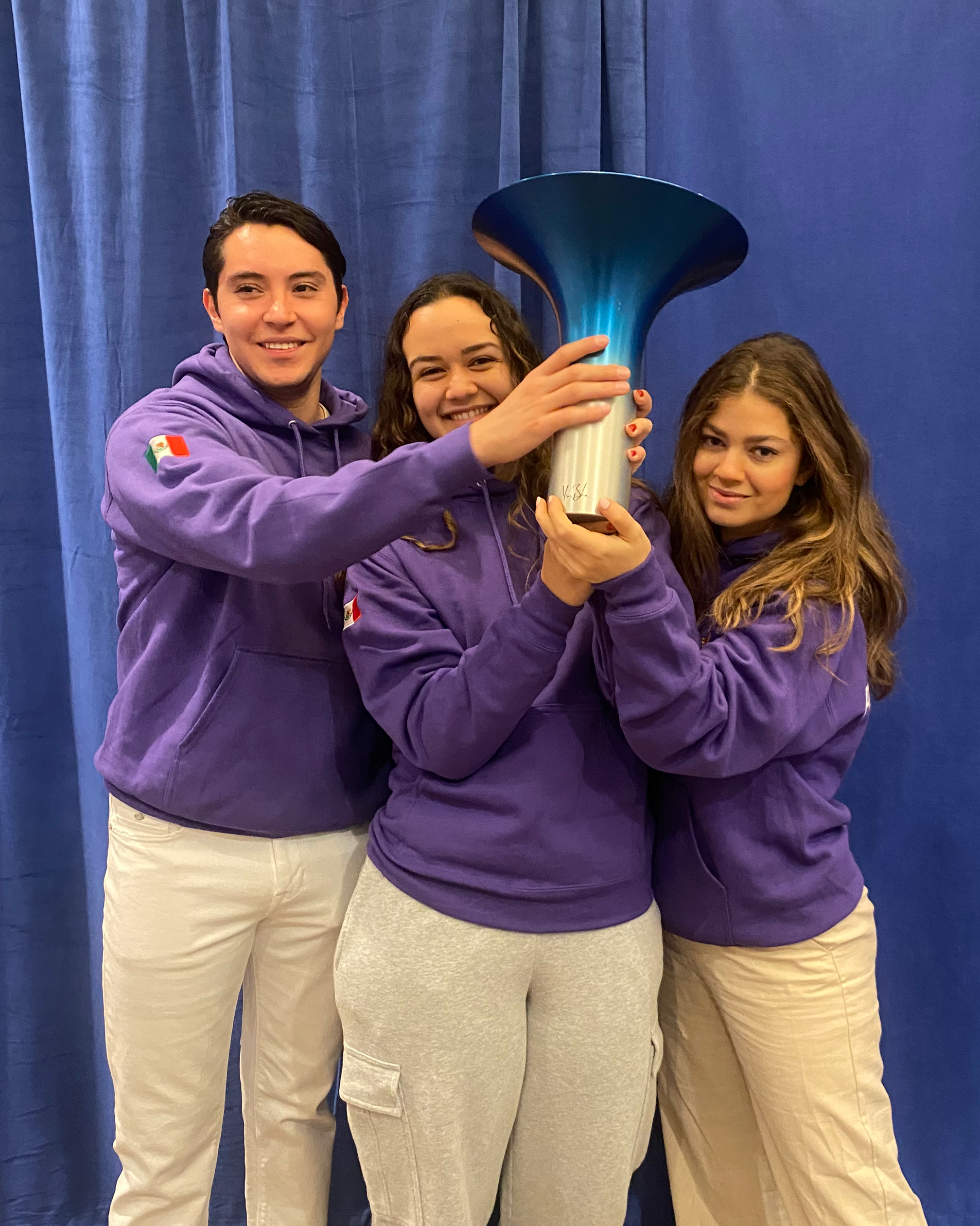 Tres jóvenes sosteniendo juntos un trofeo de forma de campana en un escenario con cortinas azules.
